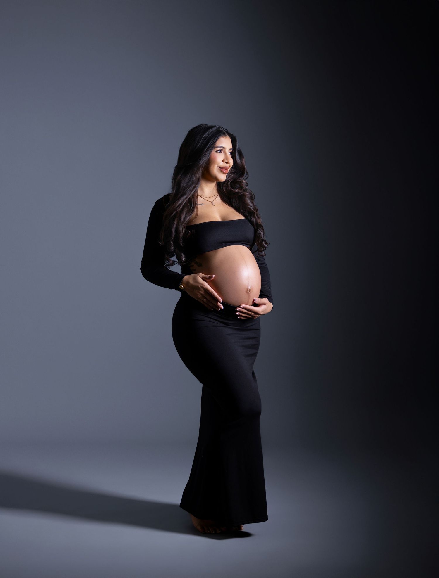 Pregnant woman in black off-shoulder crop top and skirt cradling baby bump in professional maternity photo studio.