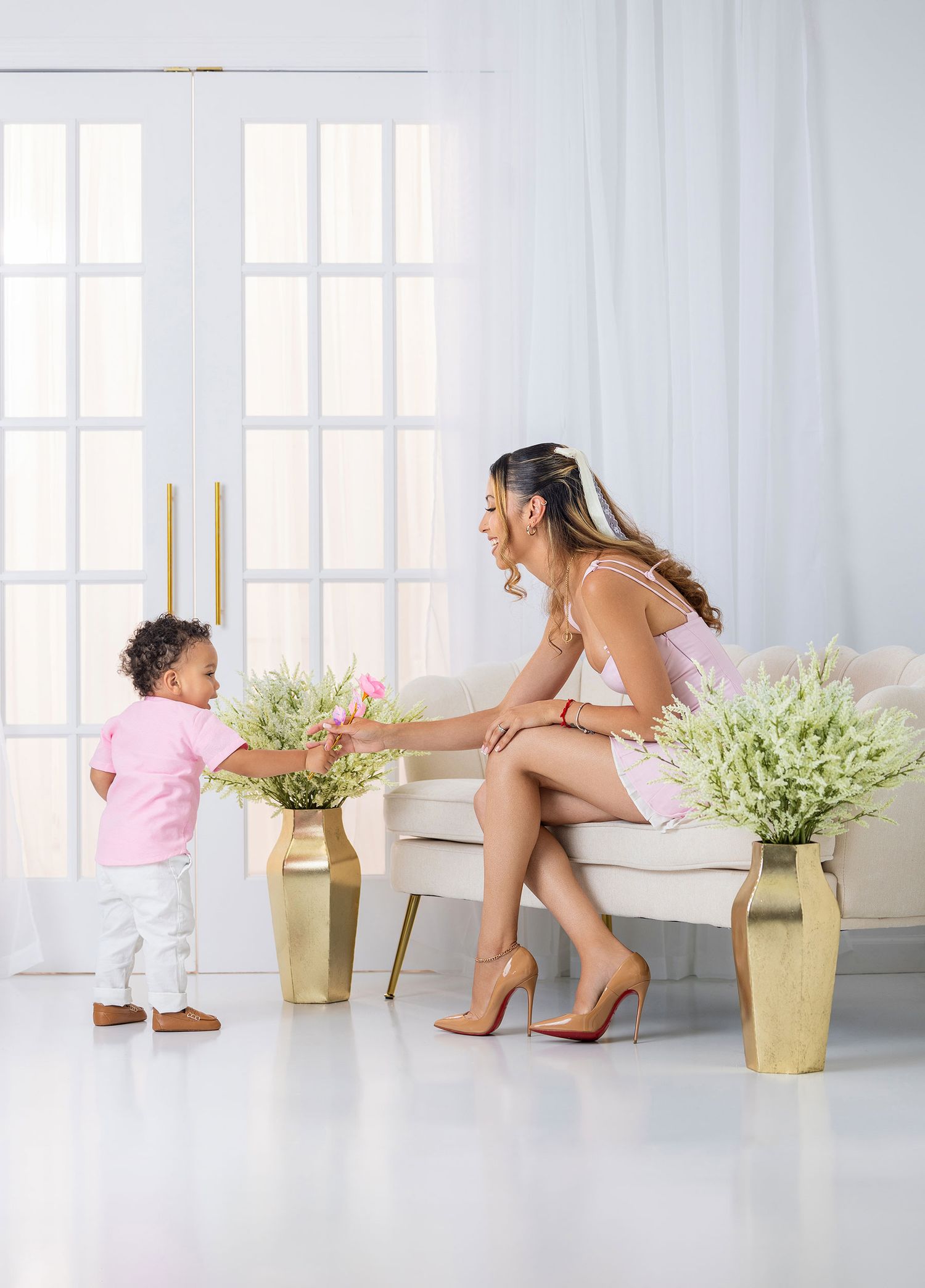 Woman in pink outfit seated on cream sofa as toddler offers flowers in bright white room.