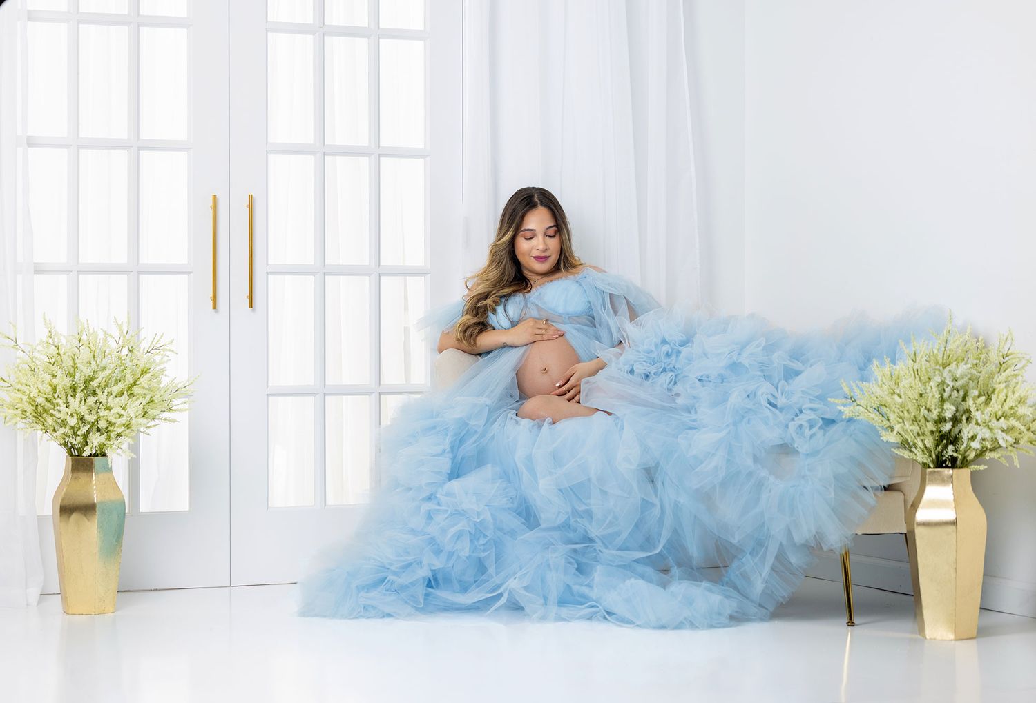 Person in flowing light blue tulle dress posing elegantly on cream sofa in bright modern interior setting.