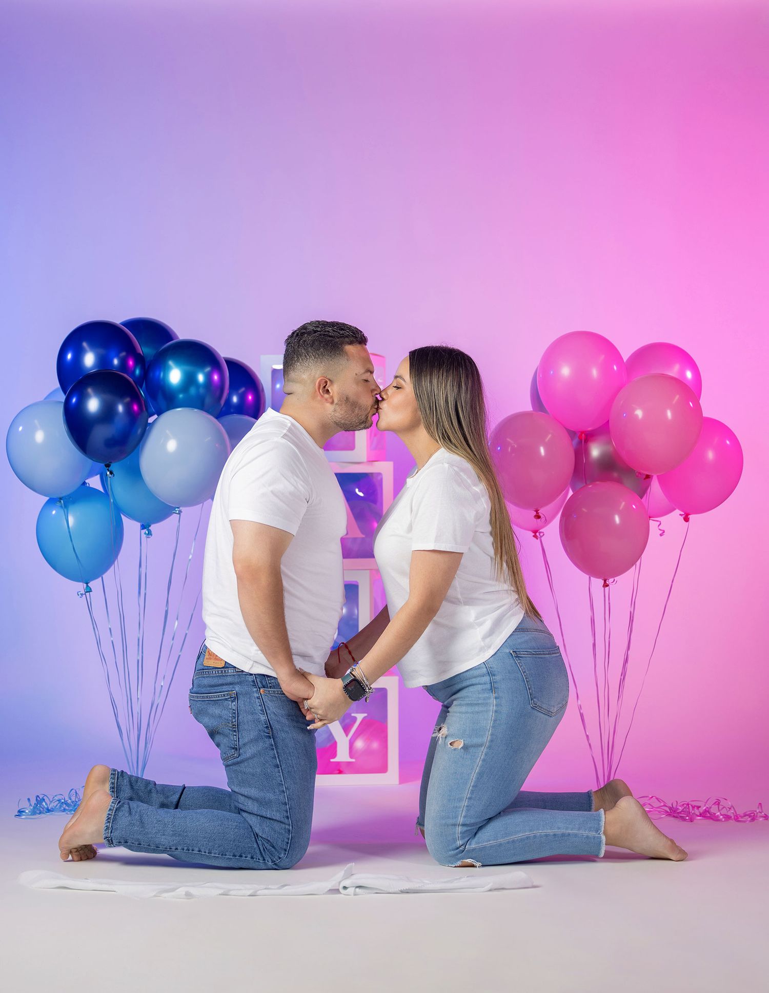 Couple kissing on knees between blue and pink balloons at gender reveal.