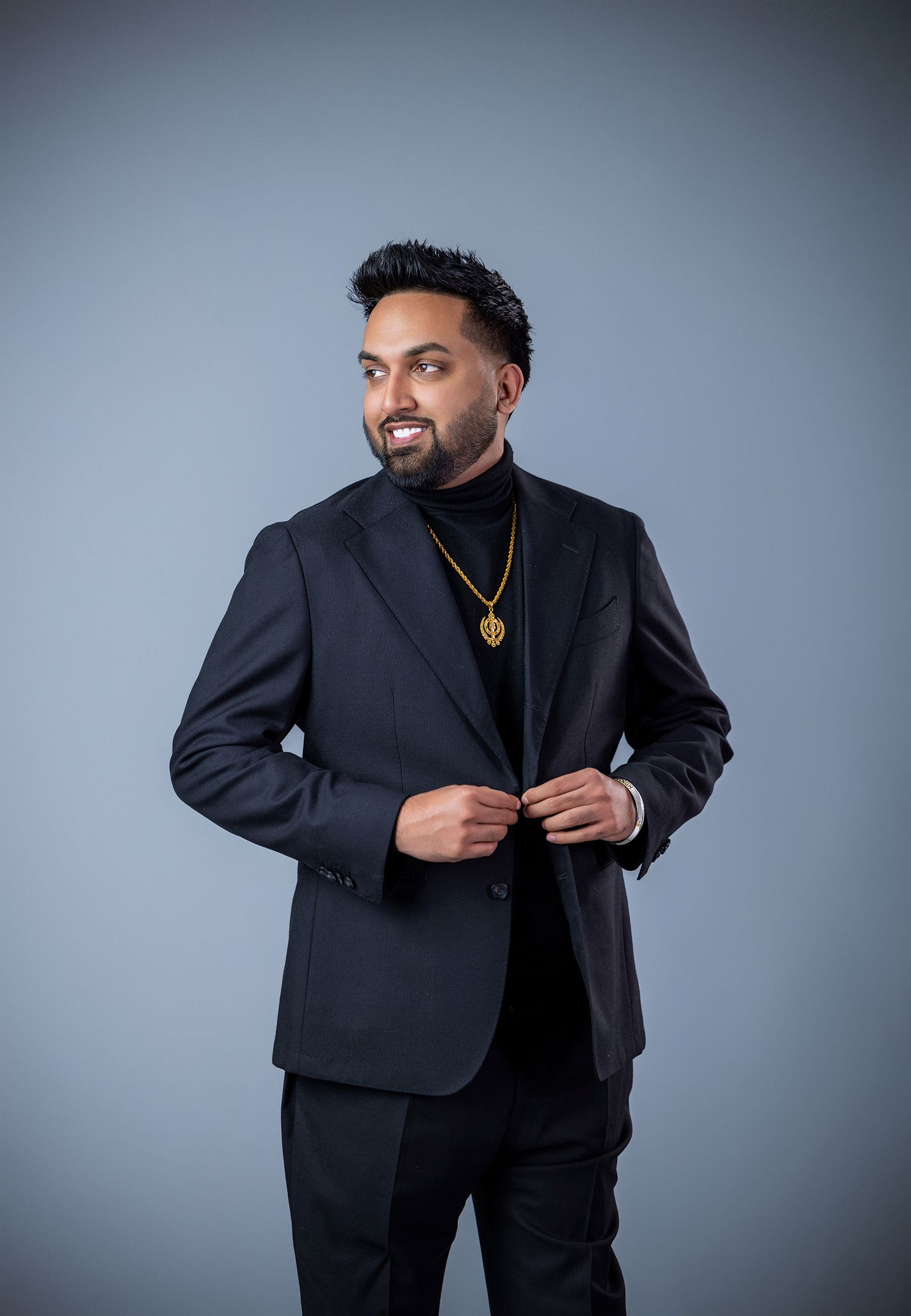 A stylish man in a black suit and turtleneck with a gold chain poses confidently against a gray background.