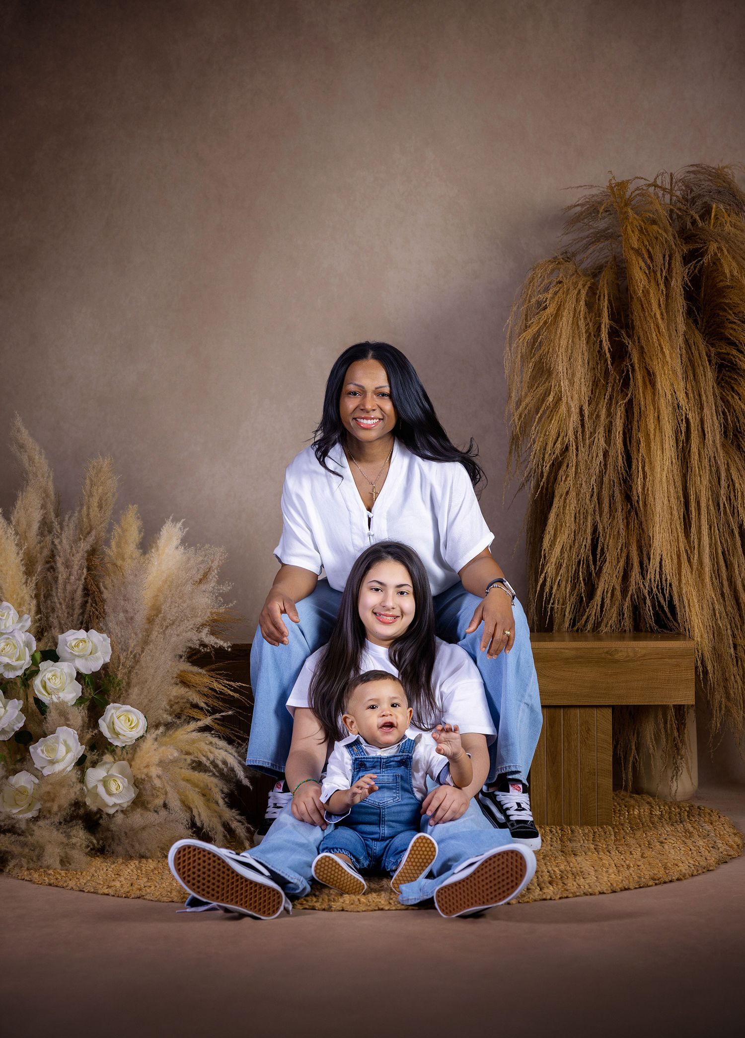 Studio portrait of two women and a baby in denim outfits, posed with dried pampas grass and white roses backdrop.