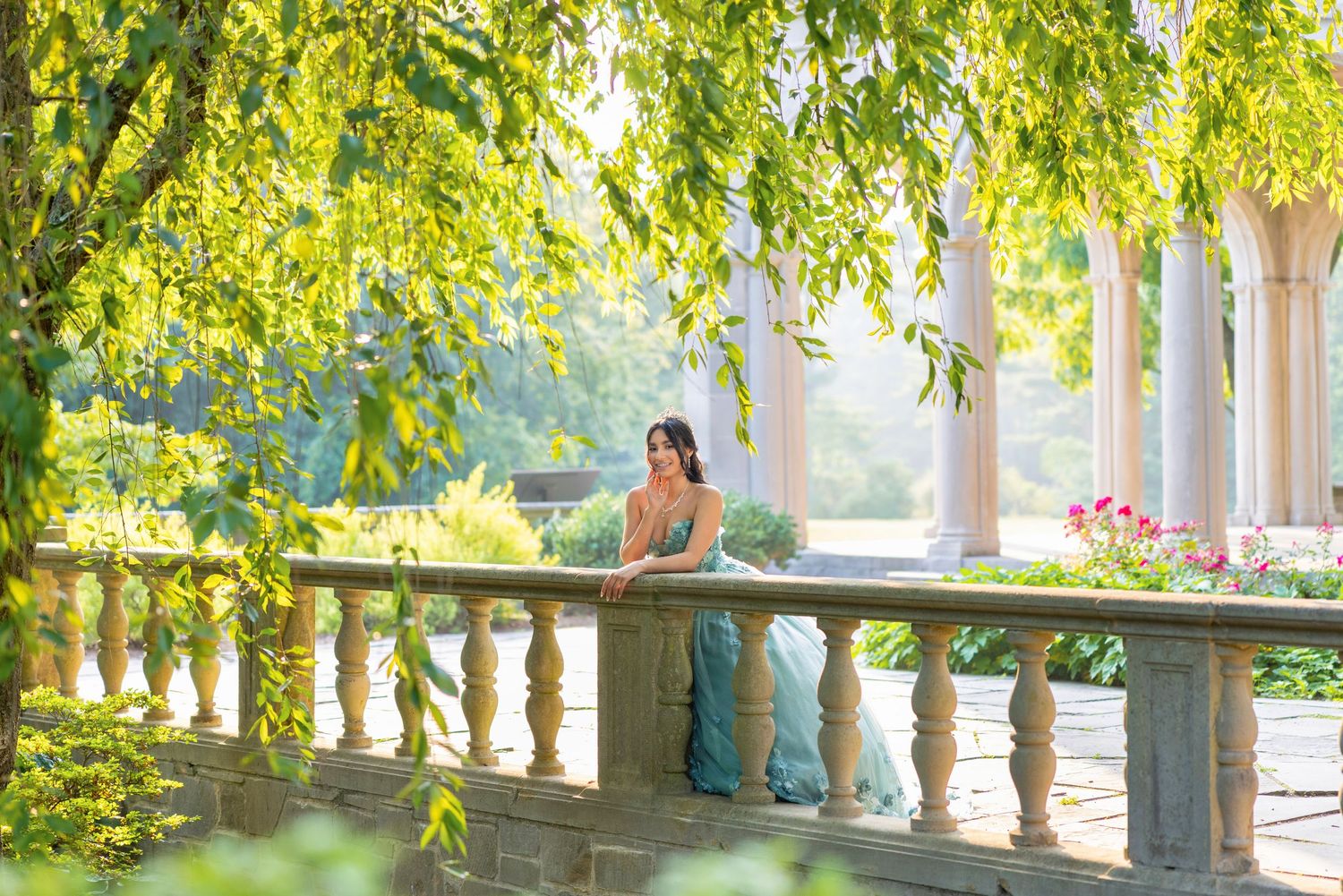 Woman in elegant gown leaning on stone balustrade surrounded by lush green weeping willow trees in a classical garden.