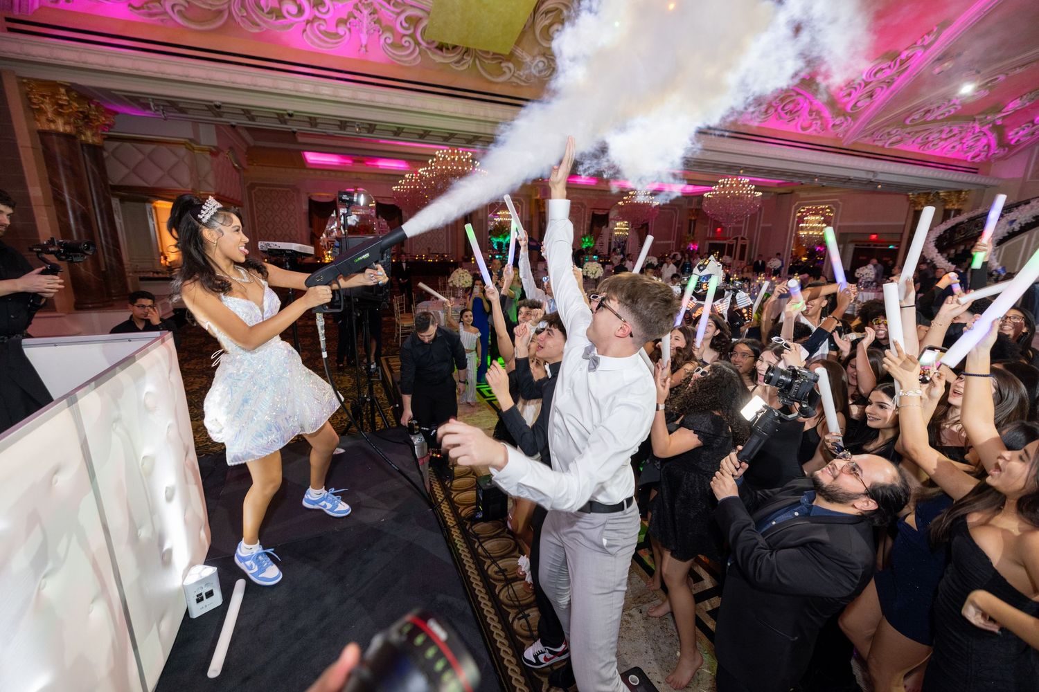 Wedding guests dancing and celebrating on a crowded dance floor with raised hands under pink and purple lighting.