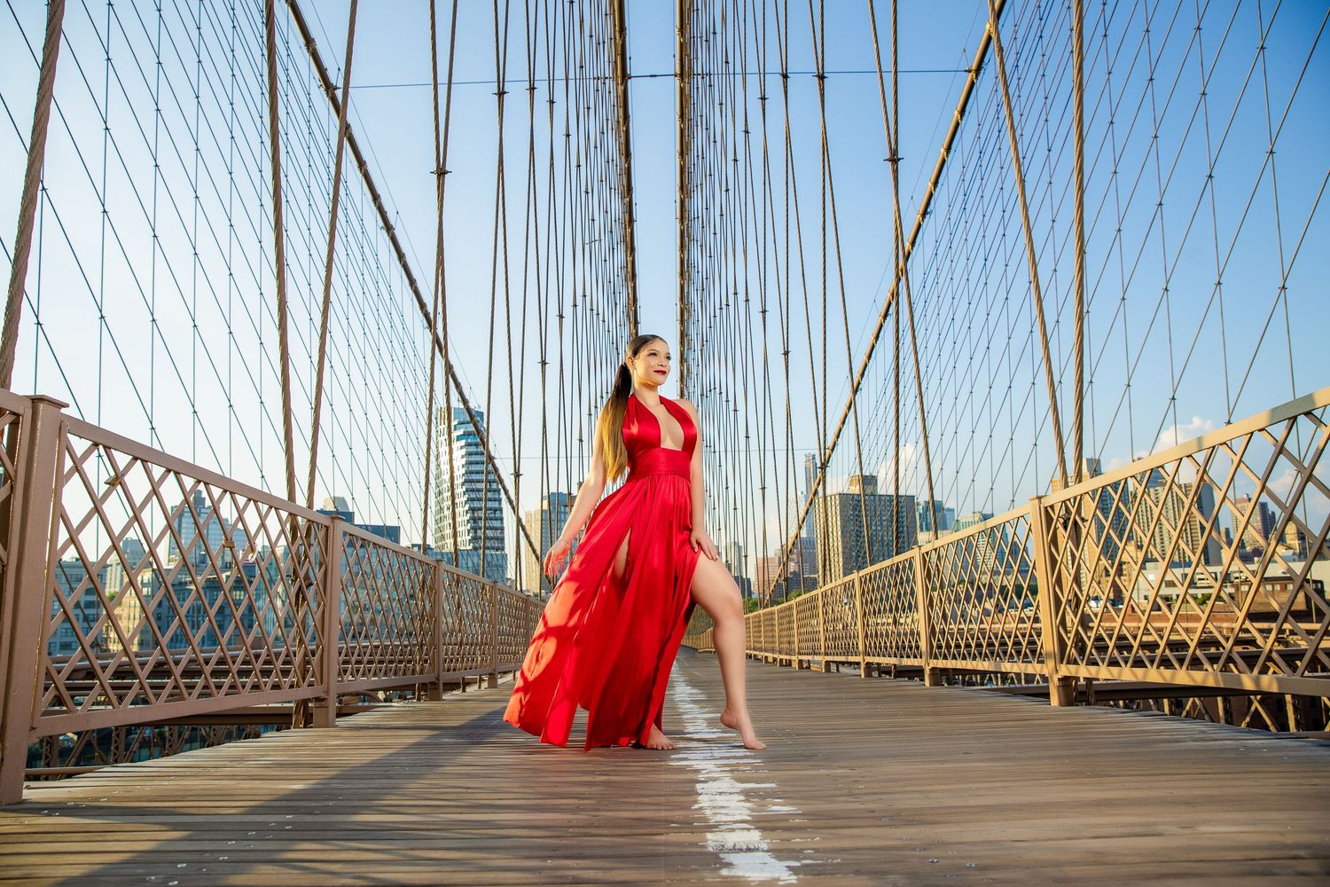 Woman in flowing red gown poses gracefully on Brooklyn Bridge walkway with NYC skyline in background.