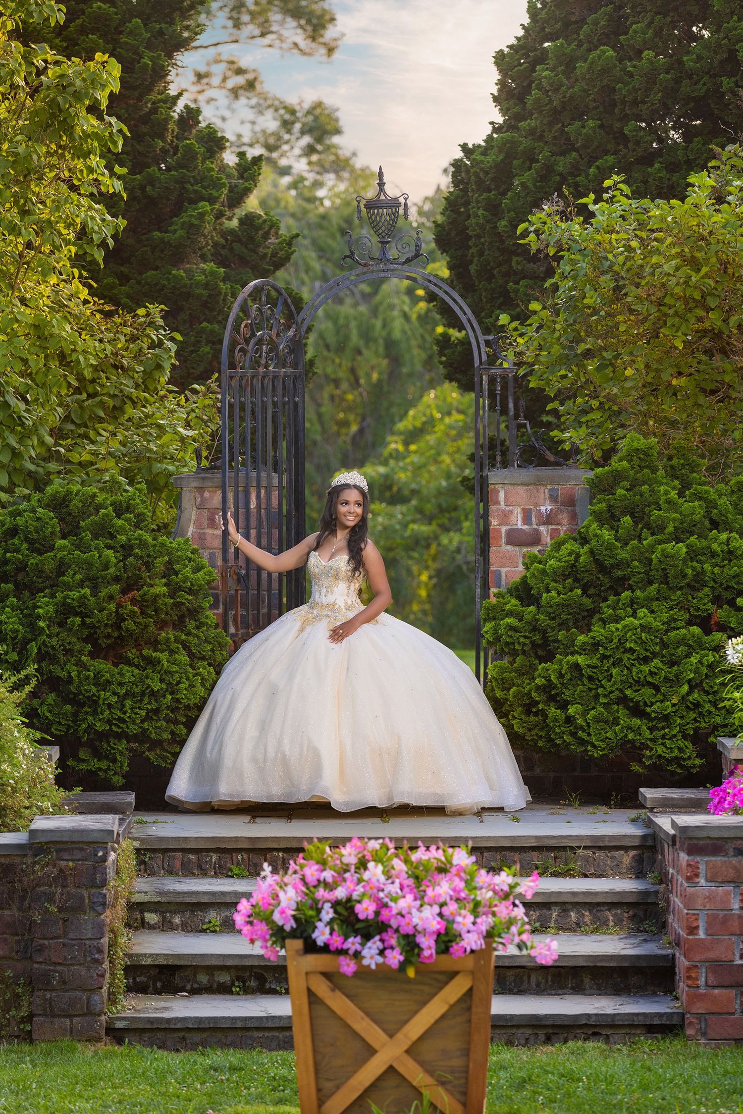 A bride in a white ballgown sits gracefully on a rustic wooden bench surrounded by lush gardens and pink flowers.