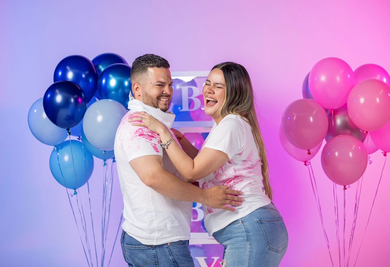 Happy couple embracing at gender reveal party with pink and blue balloon decorations.