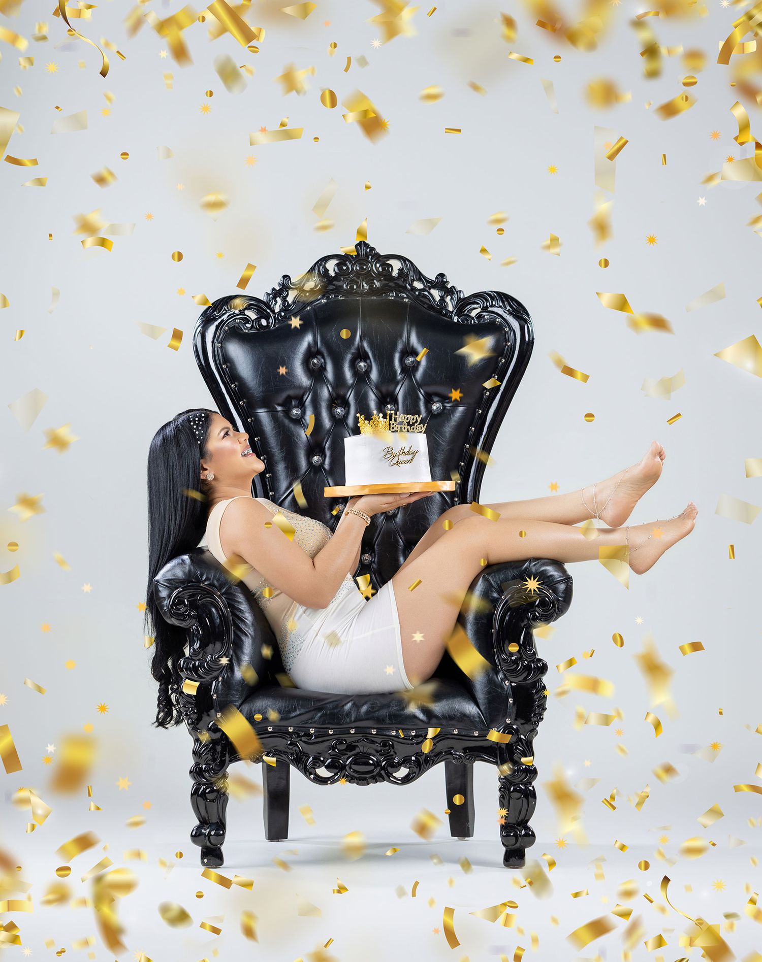 Woman in white dress on black throne chair holding birthday cake with gold confetti.