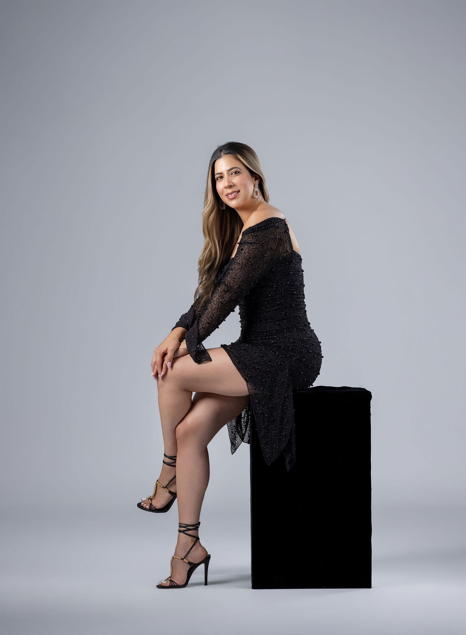 Woman in black lace mini dress and strappy heels posing seated on black pedestal against gray background.