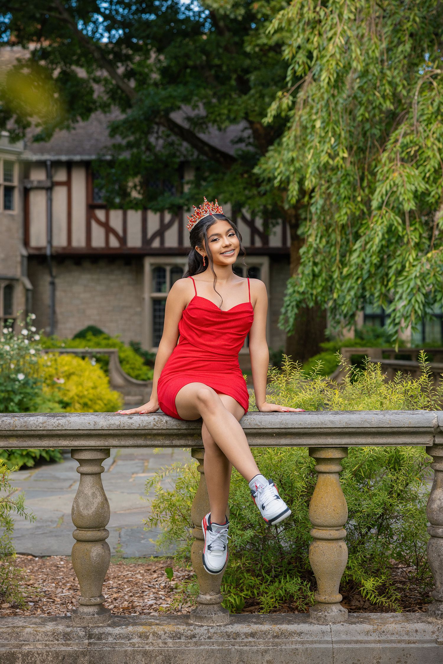 A person in a vibrant red dress sits on a stone bench in a lush garden setting with weeping willow trees.