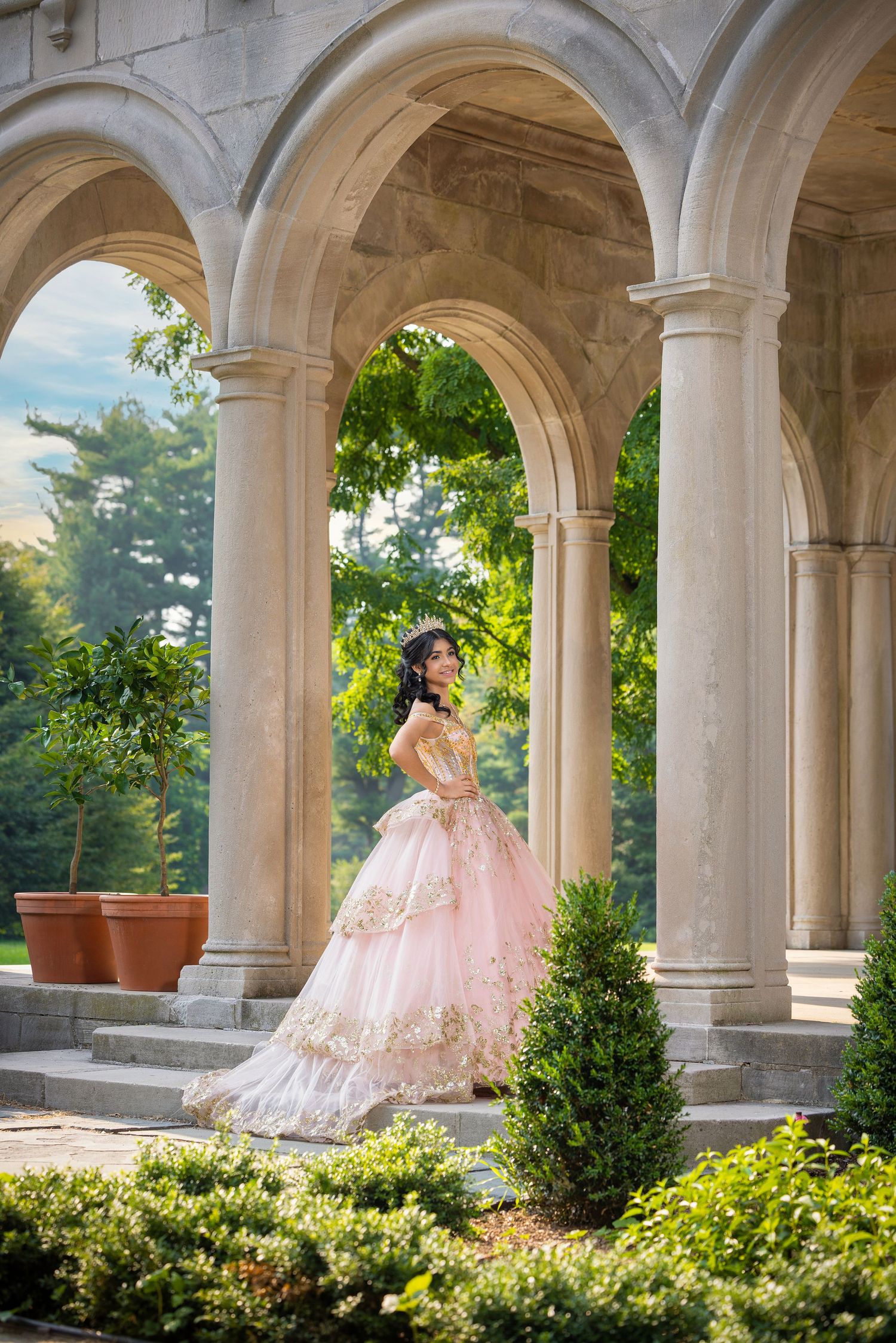 Young woman in pink gold ball gown poses gracefully under stone arches surrounded by lush greenery.