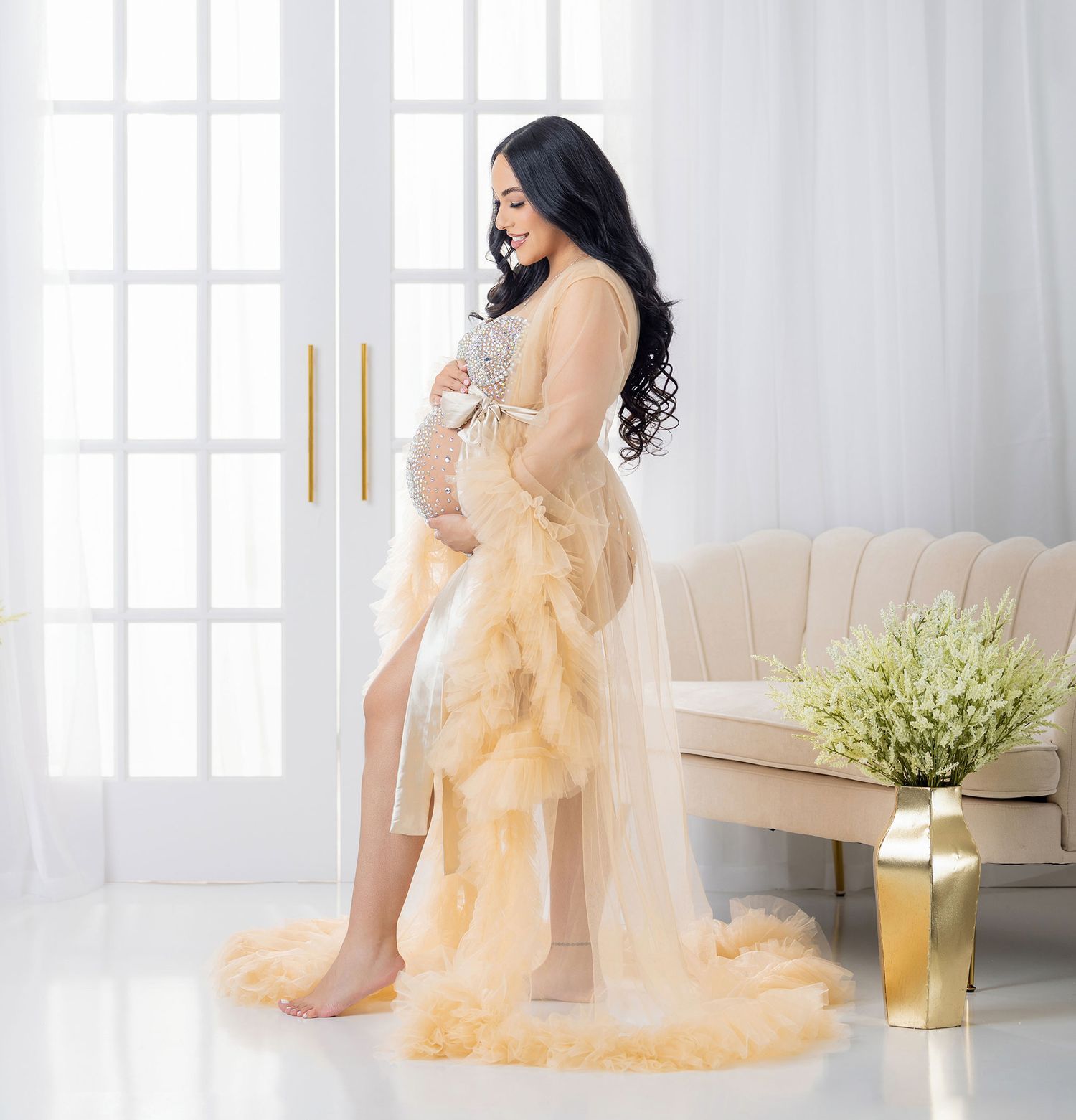 Pregnant woman in elegant champagne tulle maternity gown posing in bright white studio setting.
