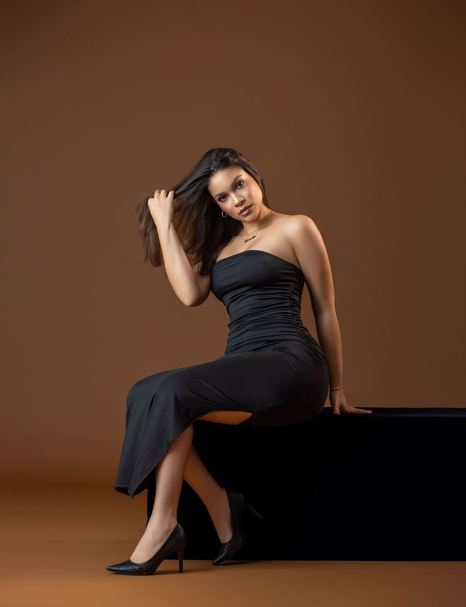 Woman in black strapless midi dress and heels poses seated against warm brown studio background.