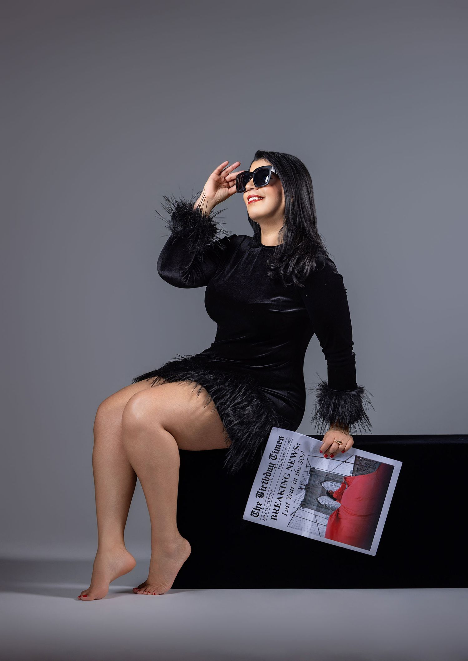 Woman in black feathered velvet dress with sunglasses seated holding a magazine in a studio.