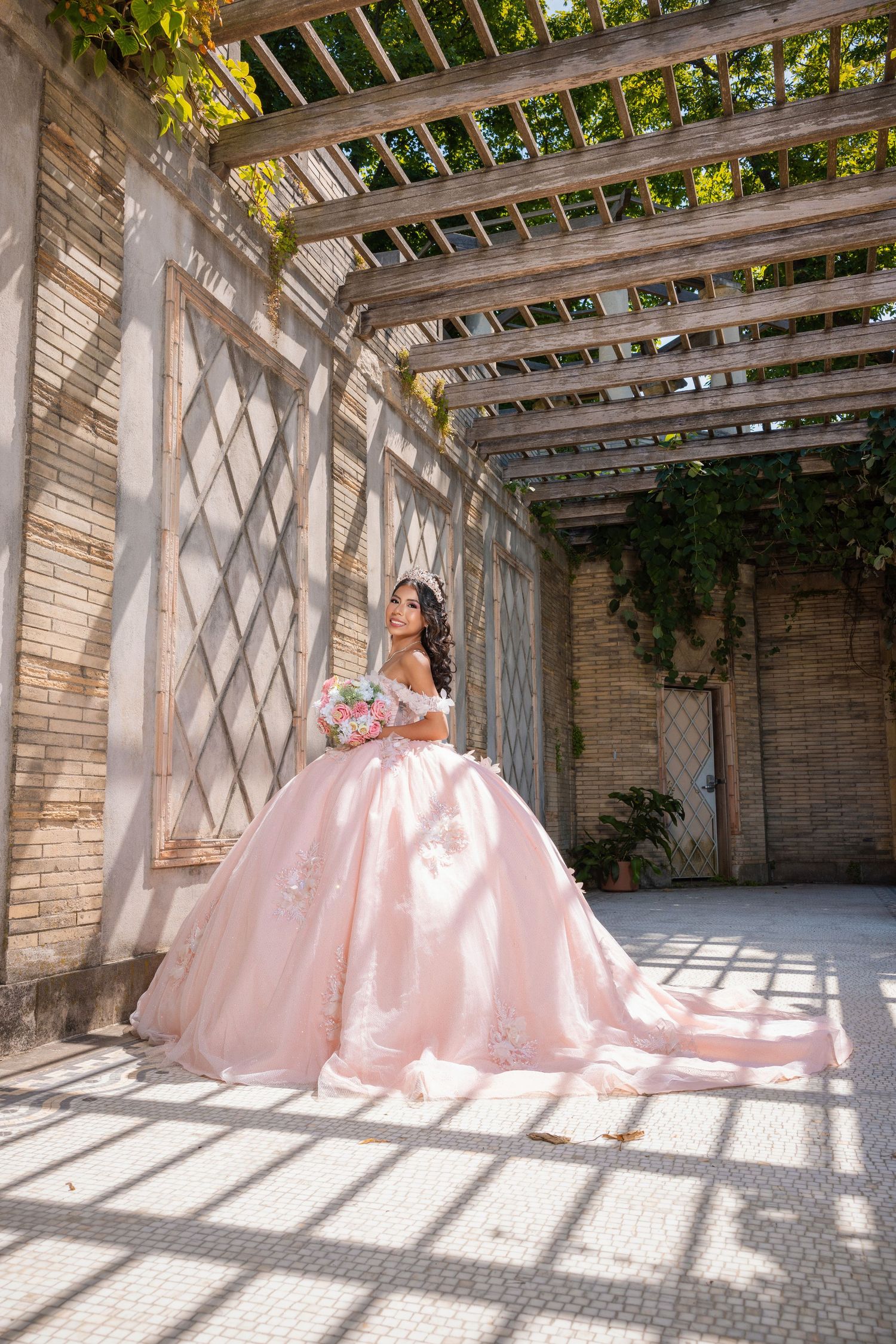 Young woman in a voluminous pink ball gown holding a bouquet under a sunlit wooden pergola with brick walls.