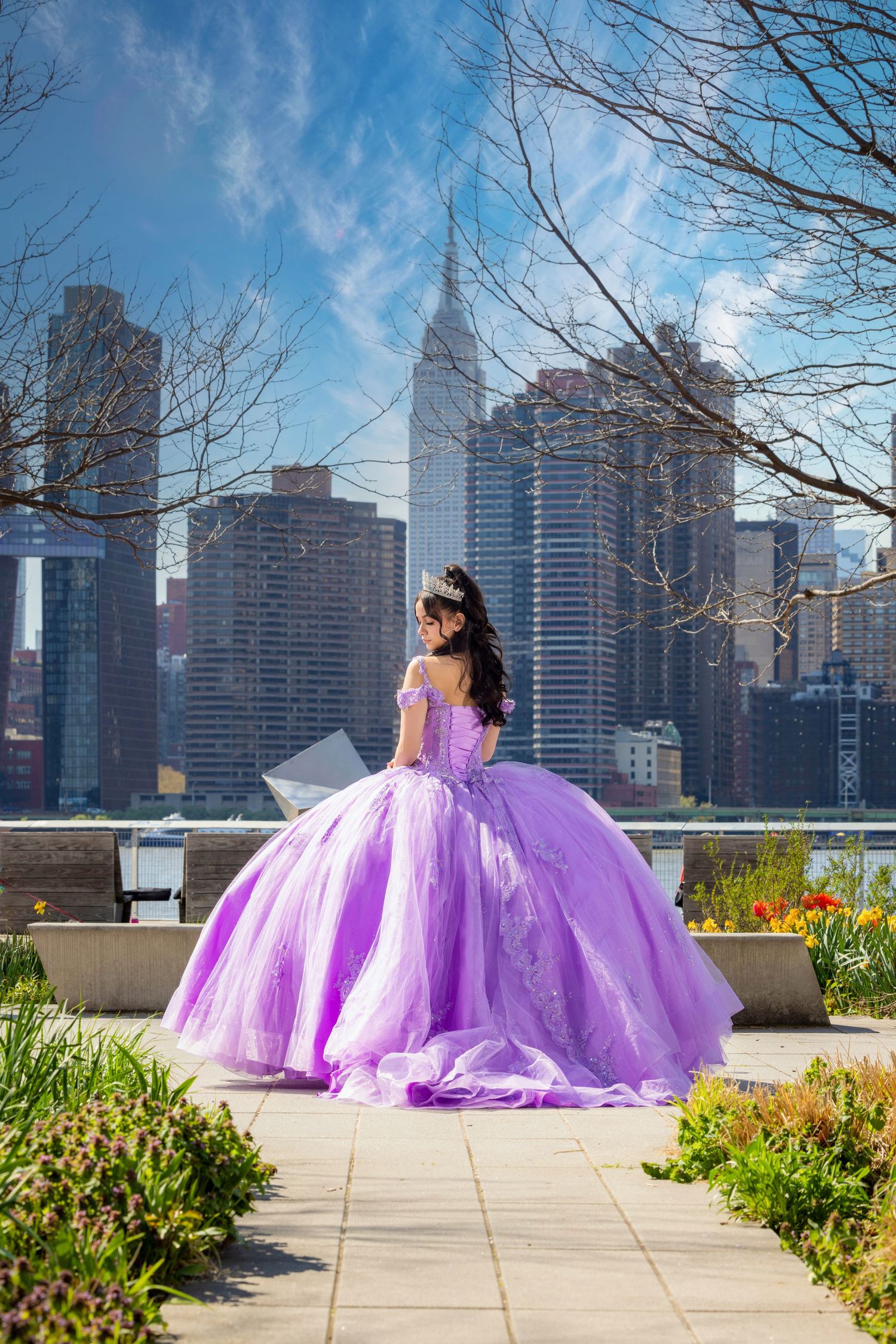 Young woman in lavish purple quinceañera ball gown poses in garden with NYC skyline backdrop on sunny day.