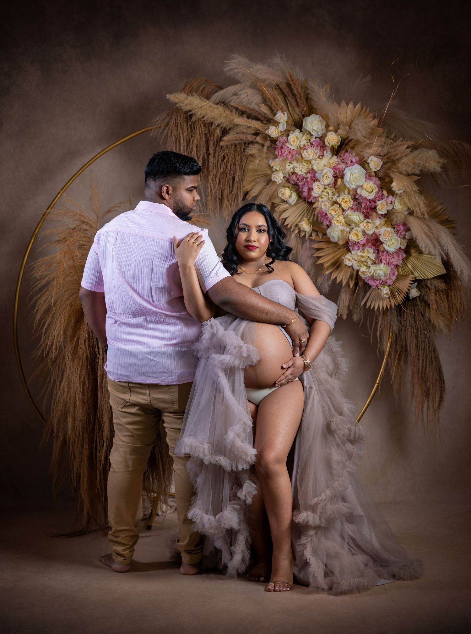A romantic maternity photoshoot featuring a couple in front of a brown backdrop with dried palm leaves and floral decor.