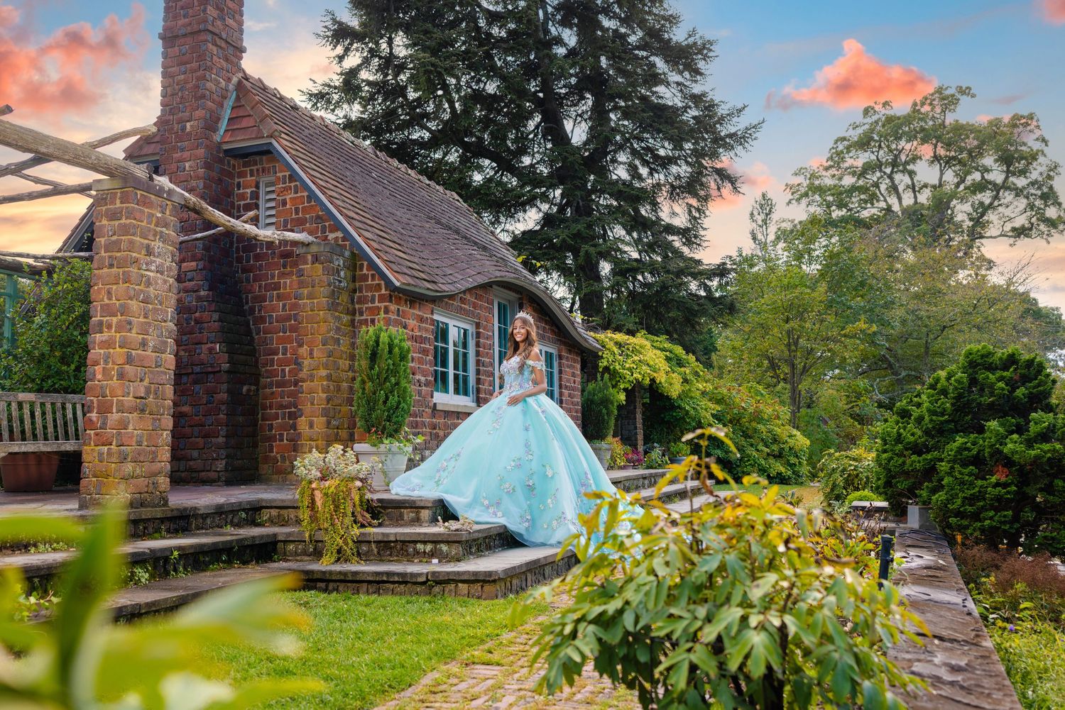Woman in elegant blue ball gown poses in lush garden outside charming brick cottage at sunset.