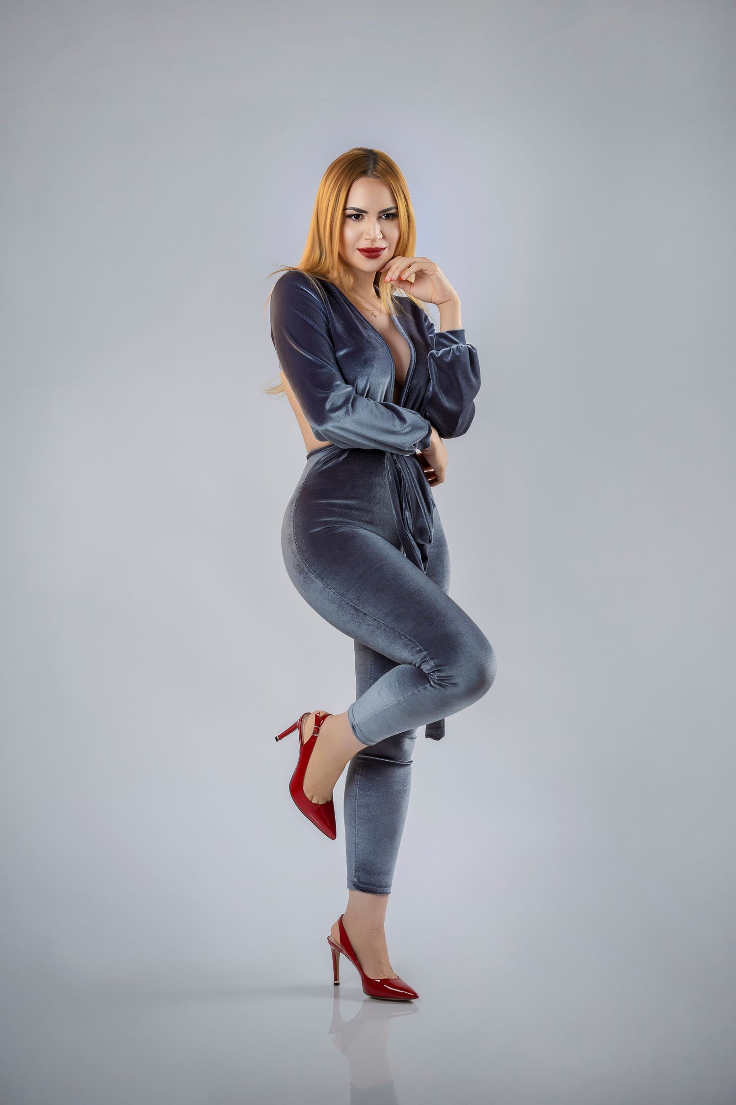 Stylish woman in gray velvet jumpsuit with red heels posing confidently in studio.