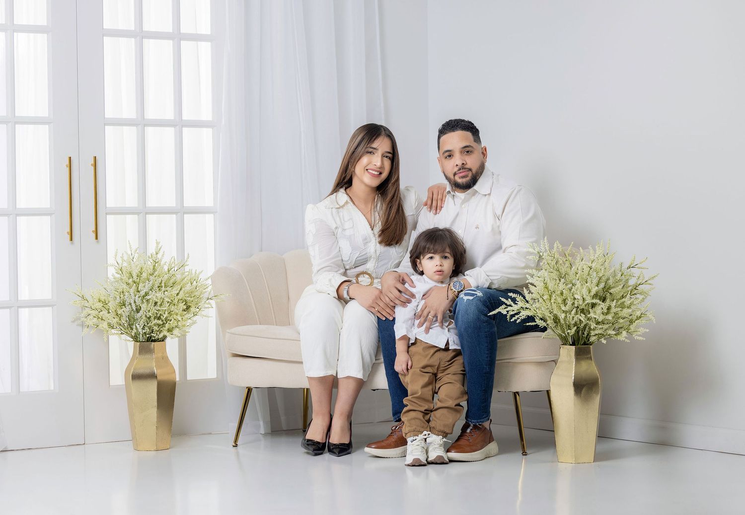 A happy family of three poses in a bright white studio, with parents seated and a toddler standing between them.