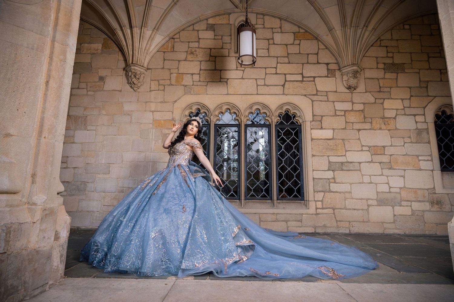 A person in a flowing blue ball gown with a dramatic train poses in a stone archway corridor.