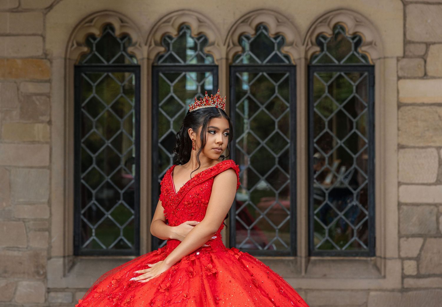A red quinceañera dress sparkles against ornate gothic windows in an elegant architectural setting.