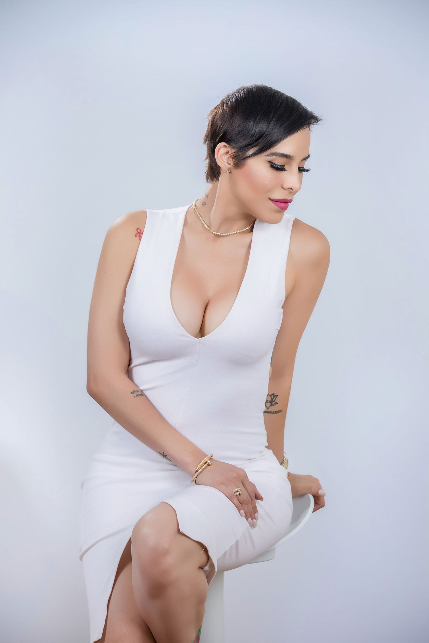 Woman in white deep-V dress with short hair, gold jewelry, and tattoos poses seated against light background.