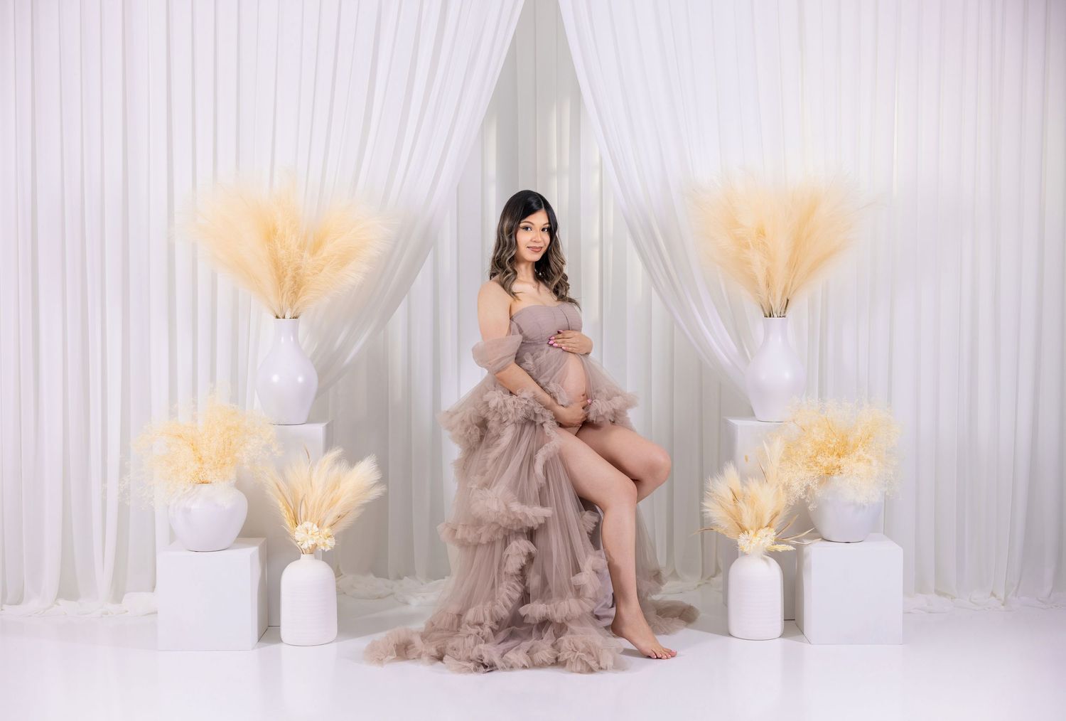 Elegant maternity photoshoot setup with flowing tulle dress and peachy pampas grass decorations against white backdrop.