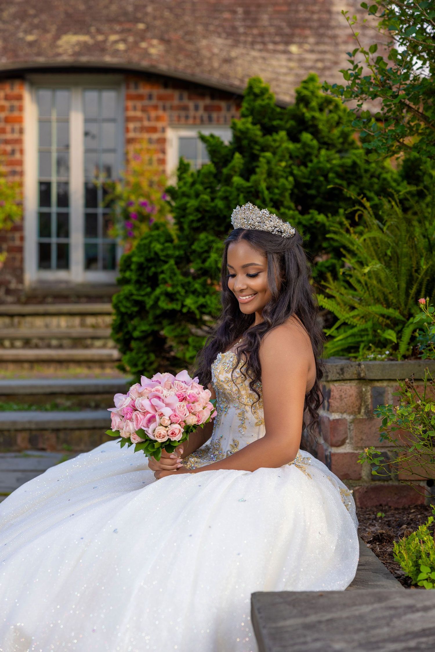 A bride in a sparkling white ballgown poses gracefully in a garden setting with pink roses and brick architecture.