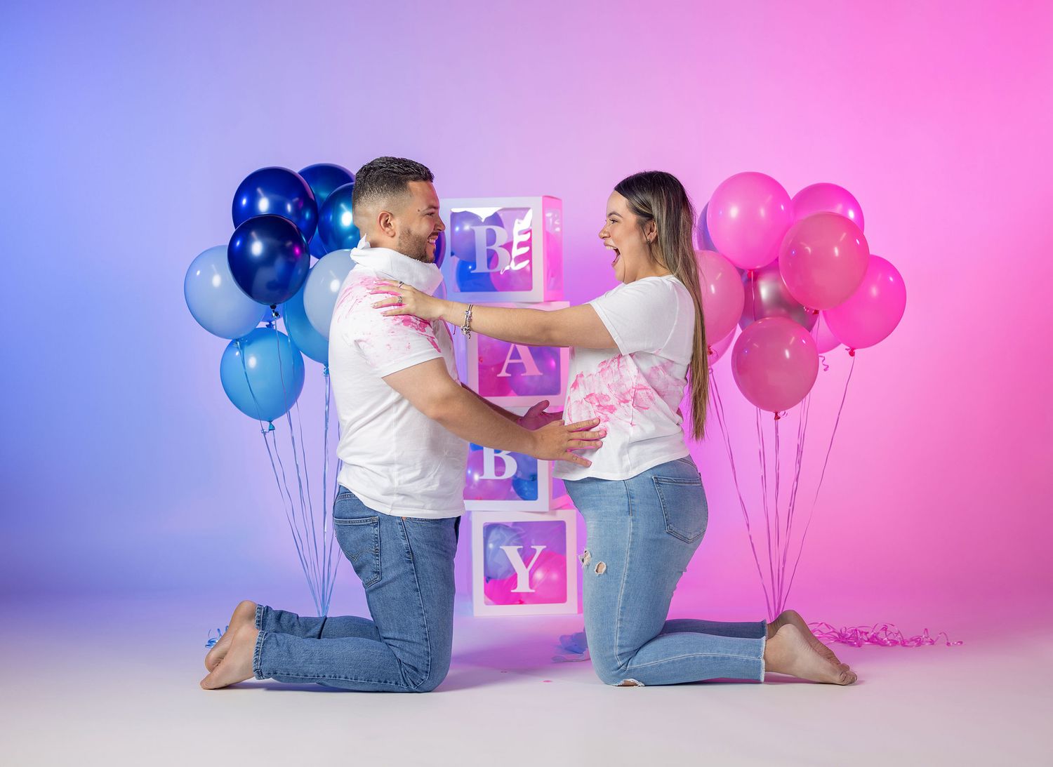 Excited couple kneeling at gender reveal with pink and blue balloons spelling BABY.