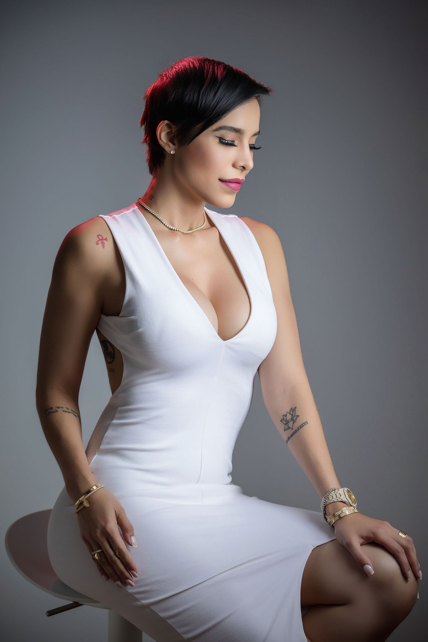 Woman in white deep-V dress with tattoos and gold jewelry seated on stool against gray background.