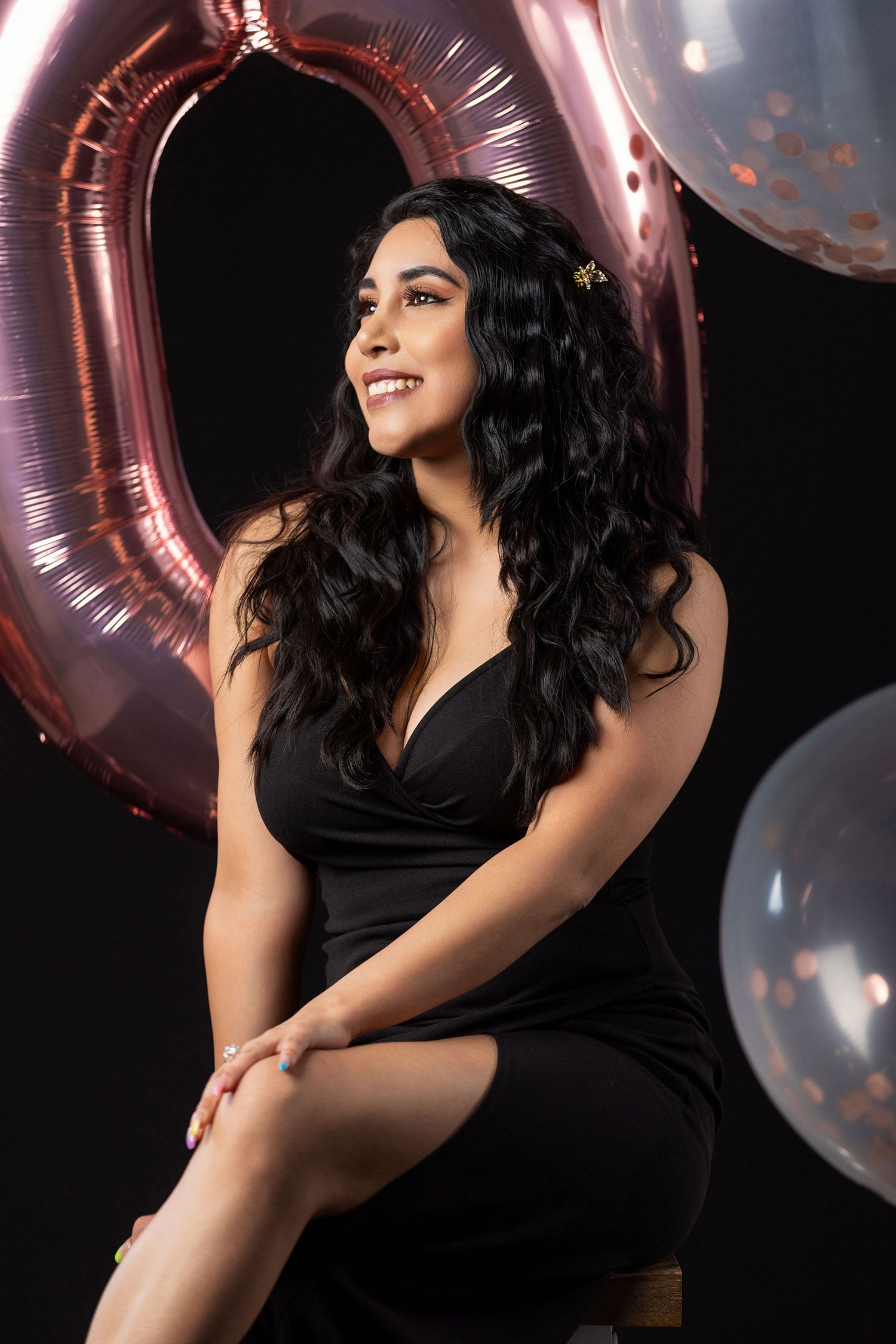 Woman in black dress smiles beside rose gold number balloon in celebration portrait.