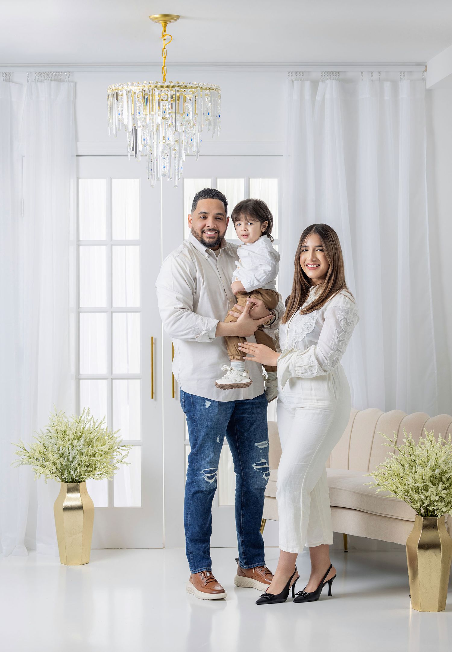 Family of three with parents holding young child posing under chandelier in white studio.
