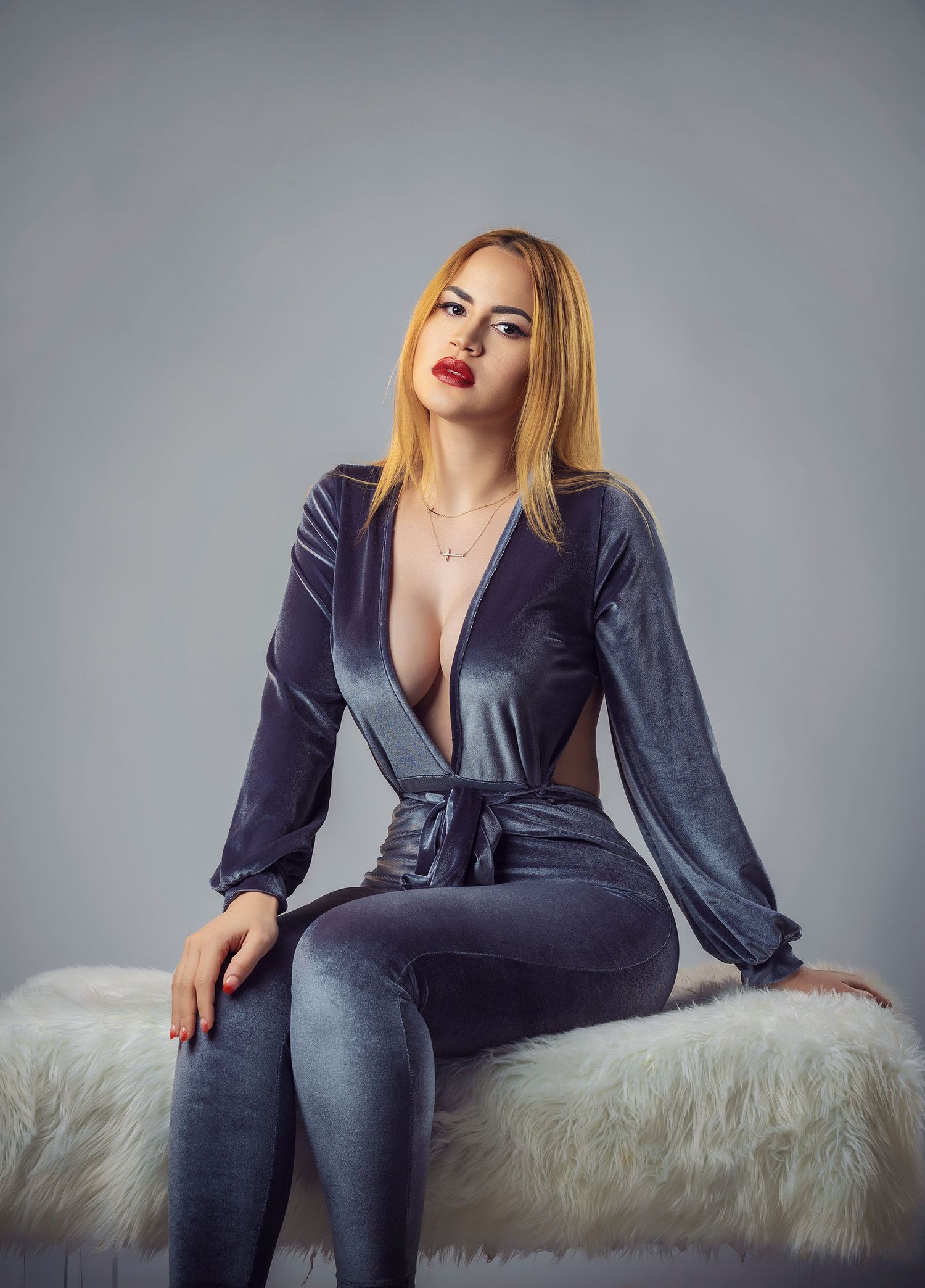 Woman in gray velvet jumpsuit with deep V-neckline sits on white fur bench, posing confidently against gray background.