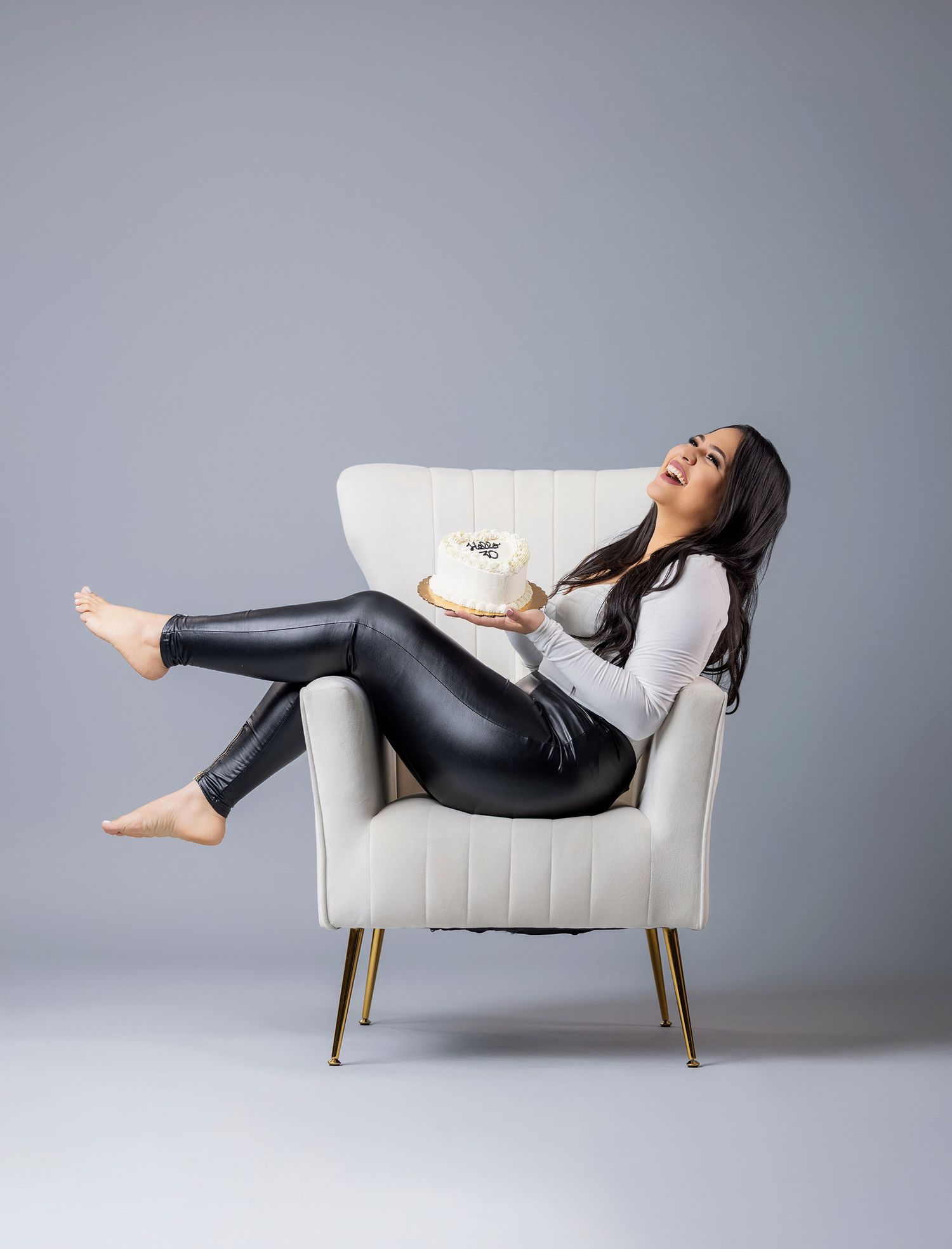 Woman in black leather pants holding a white cake while relaxing in a cream velvet accent chair with gold legs.