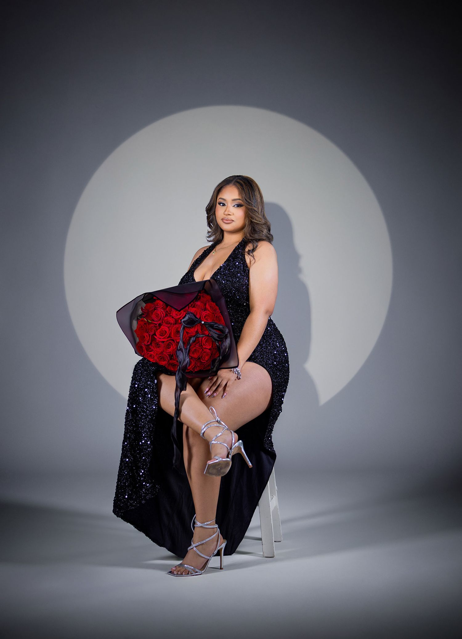 Woman in black sequin gown holding red roses bouquet, seated against spotlight backdrop wearing strappy heels.