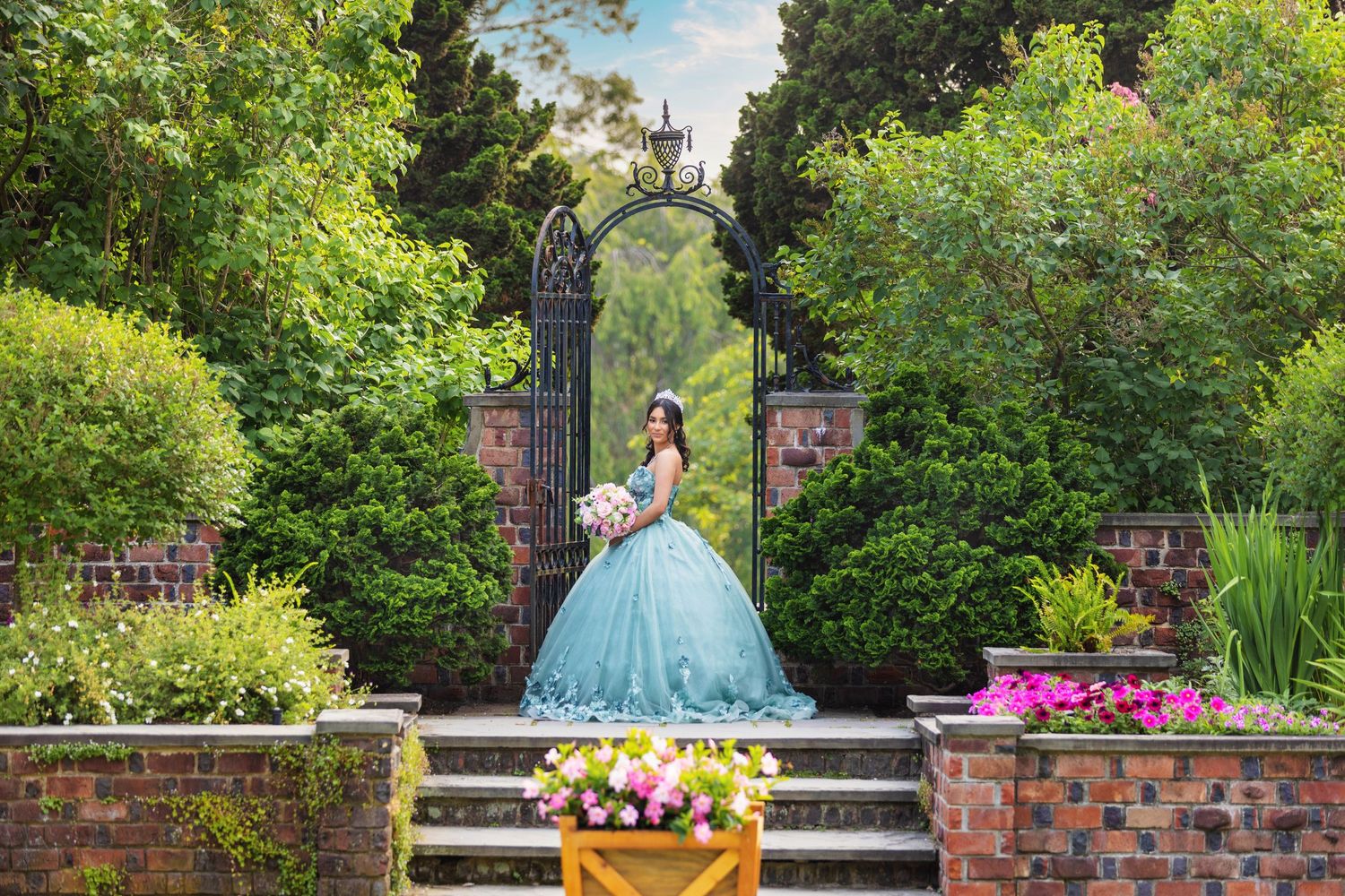 Young woman in a teal ball gown holding a bouquet stands before an ornate garden gate surrounded by lush greenery.