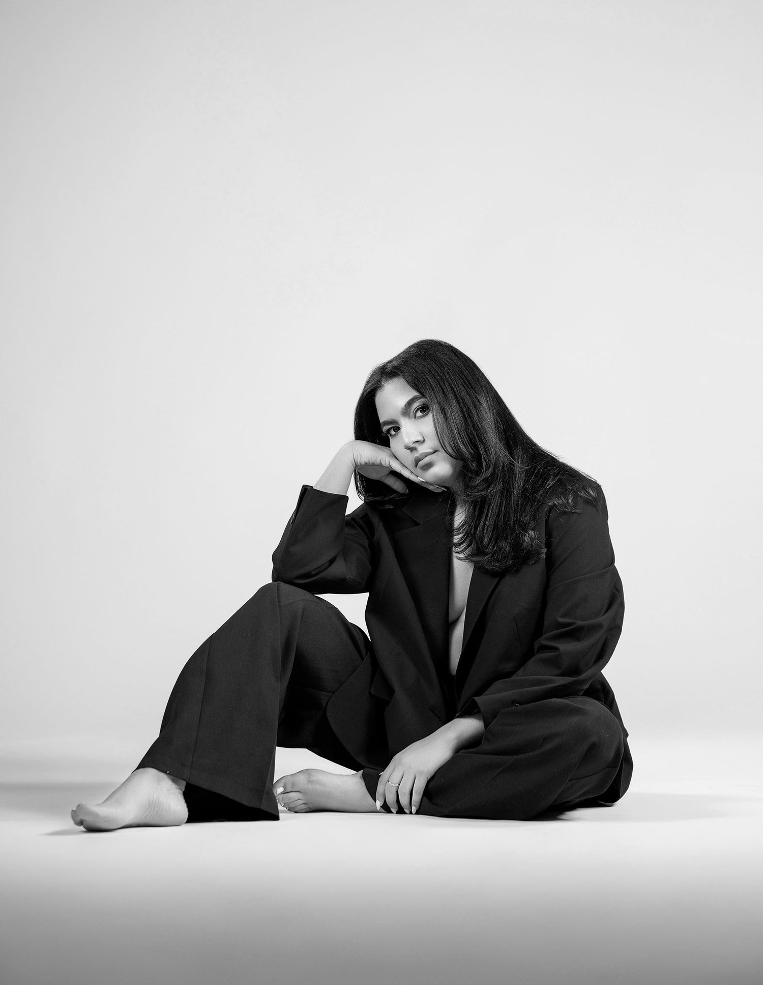 Woman in black blazer and trousers sitting barefoot on floor in black and white portrait photo.
