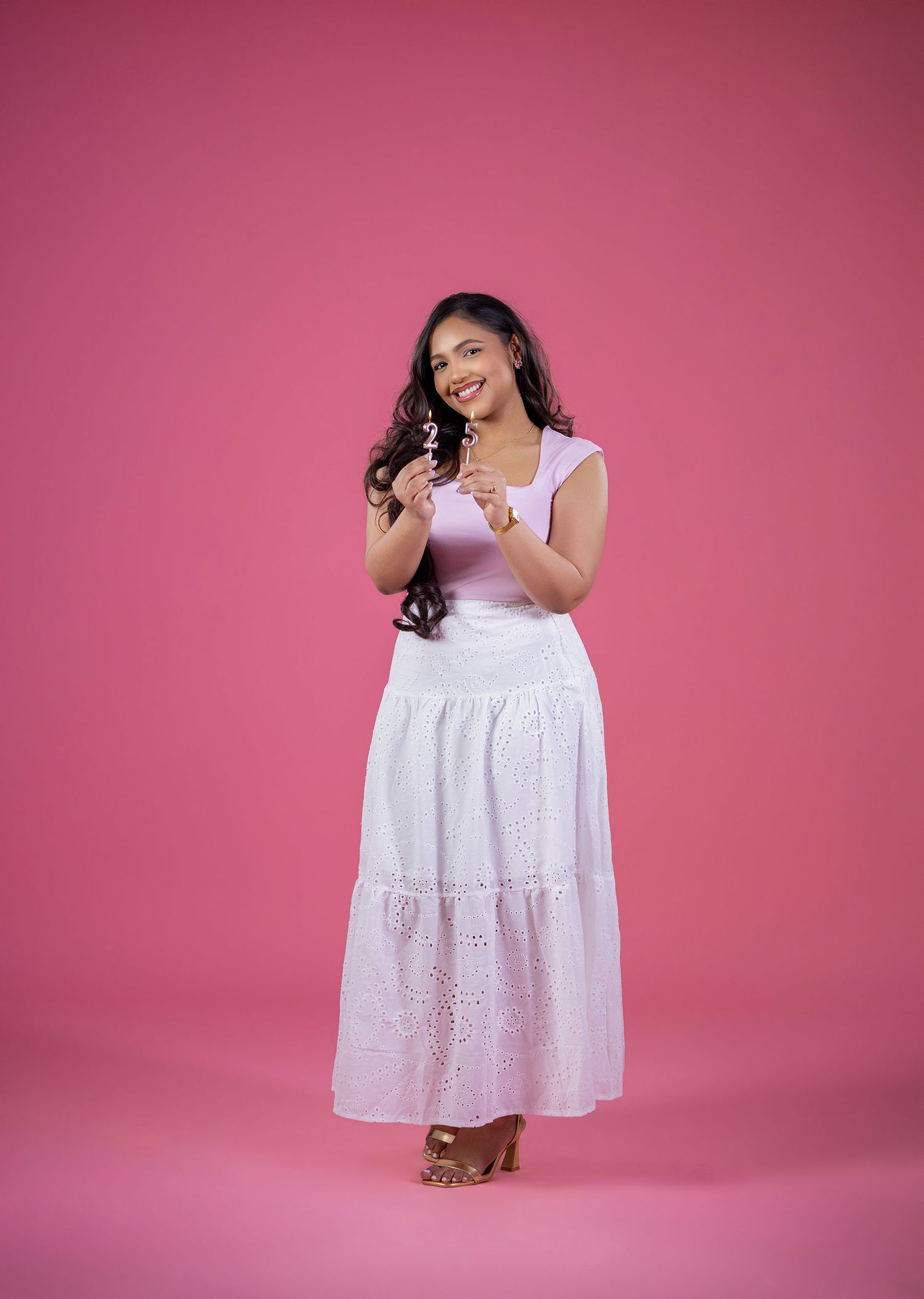 Young woman in white tiered maxi skirt and pink top smiling on pink studio background.