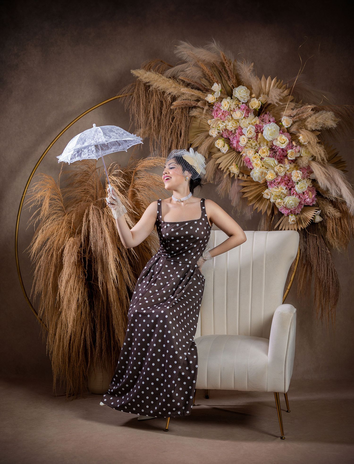 Woman in polka dot dress holding white parasol beside floral wreath in styled studio.