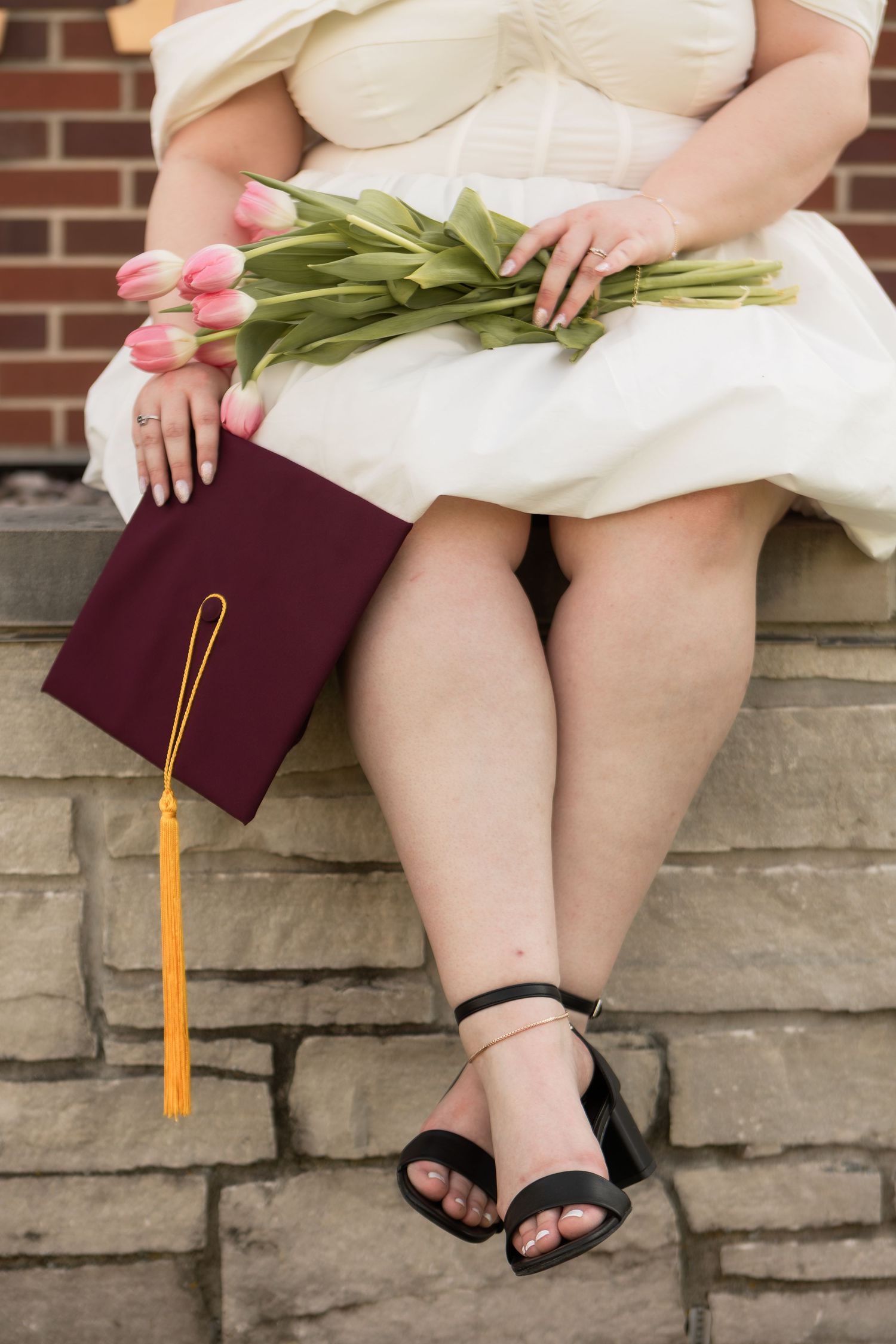 Woman sitting on stone wall in heels and white dress holding tulips and graduation cap in Columbus Ohio