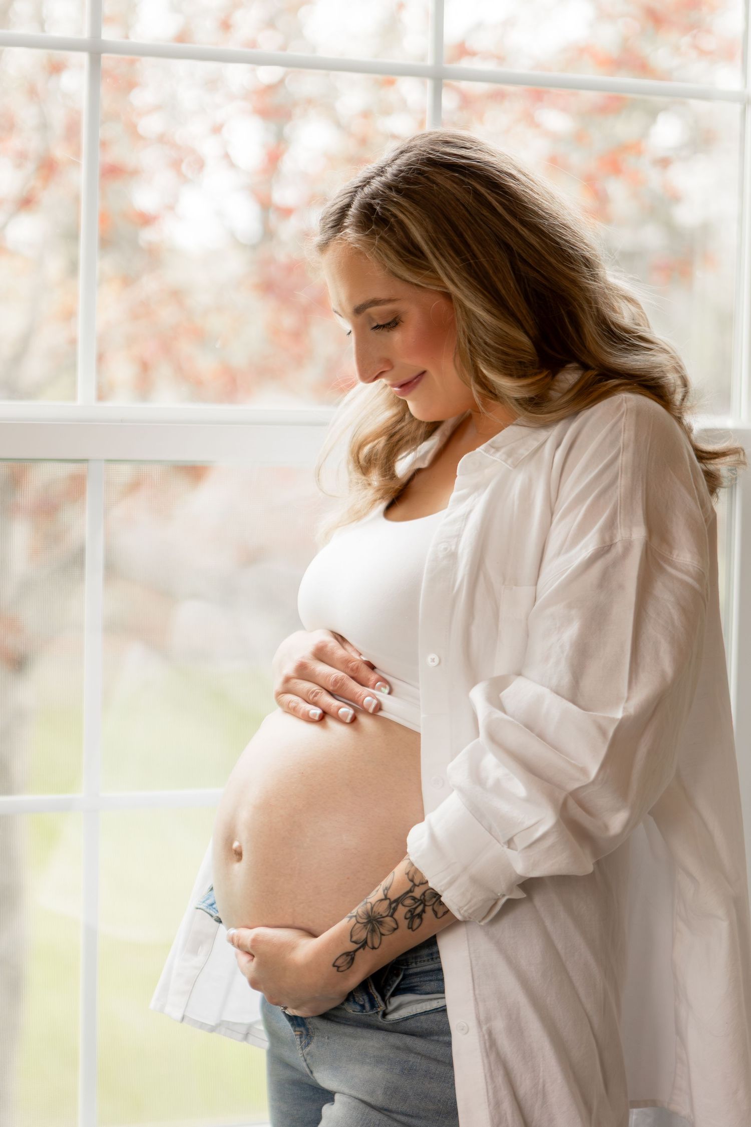 Mother standing in front of window embracing baby bump in white button down shirt and jeans in Columbus Ohio