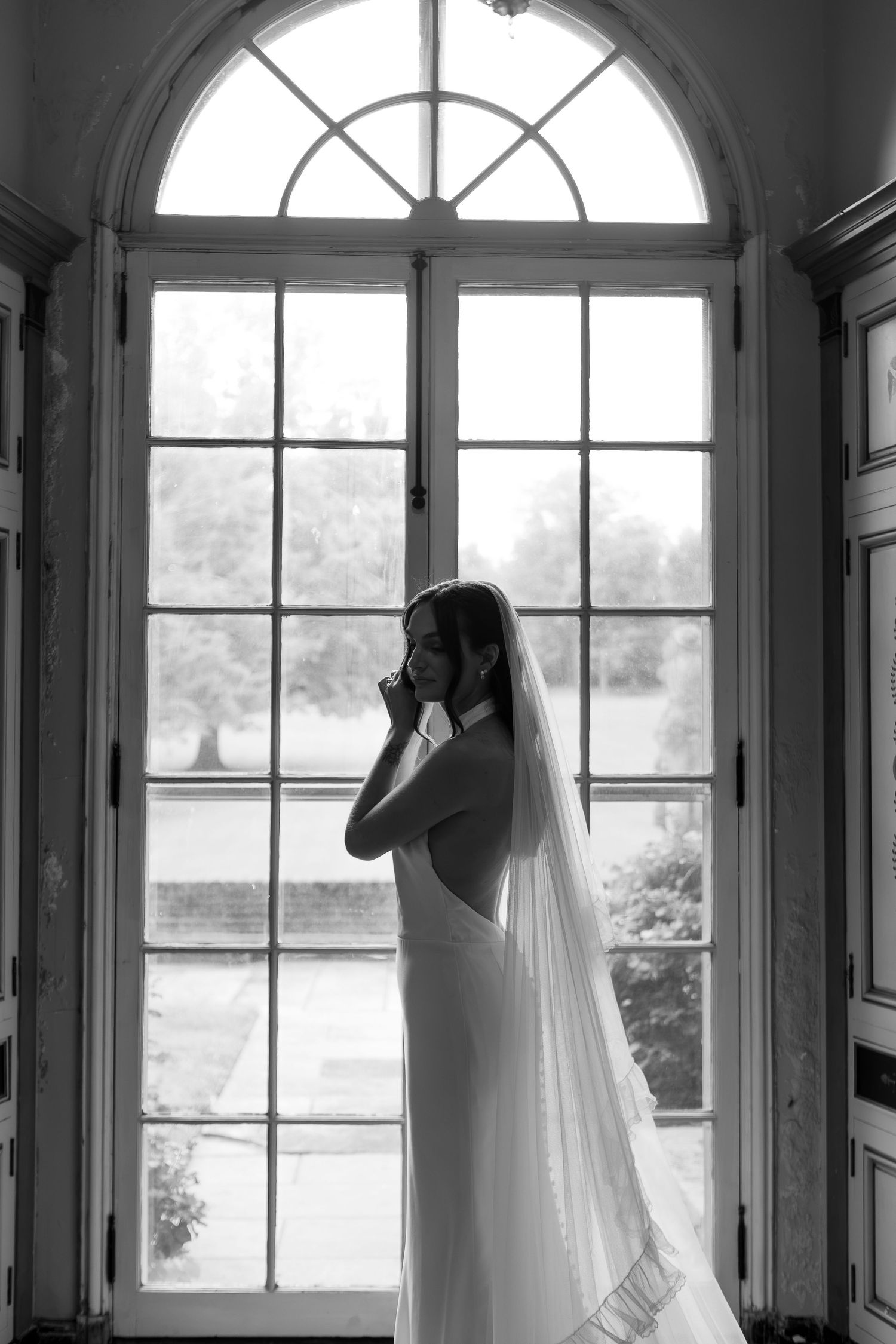 Black and white photo of a bride standing in front of large glass window fixing her earrings in Mansfield Ohio