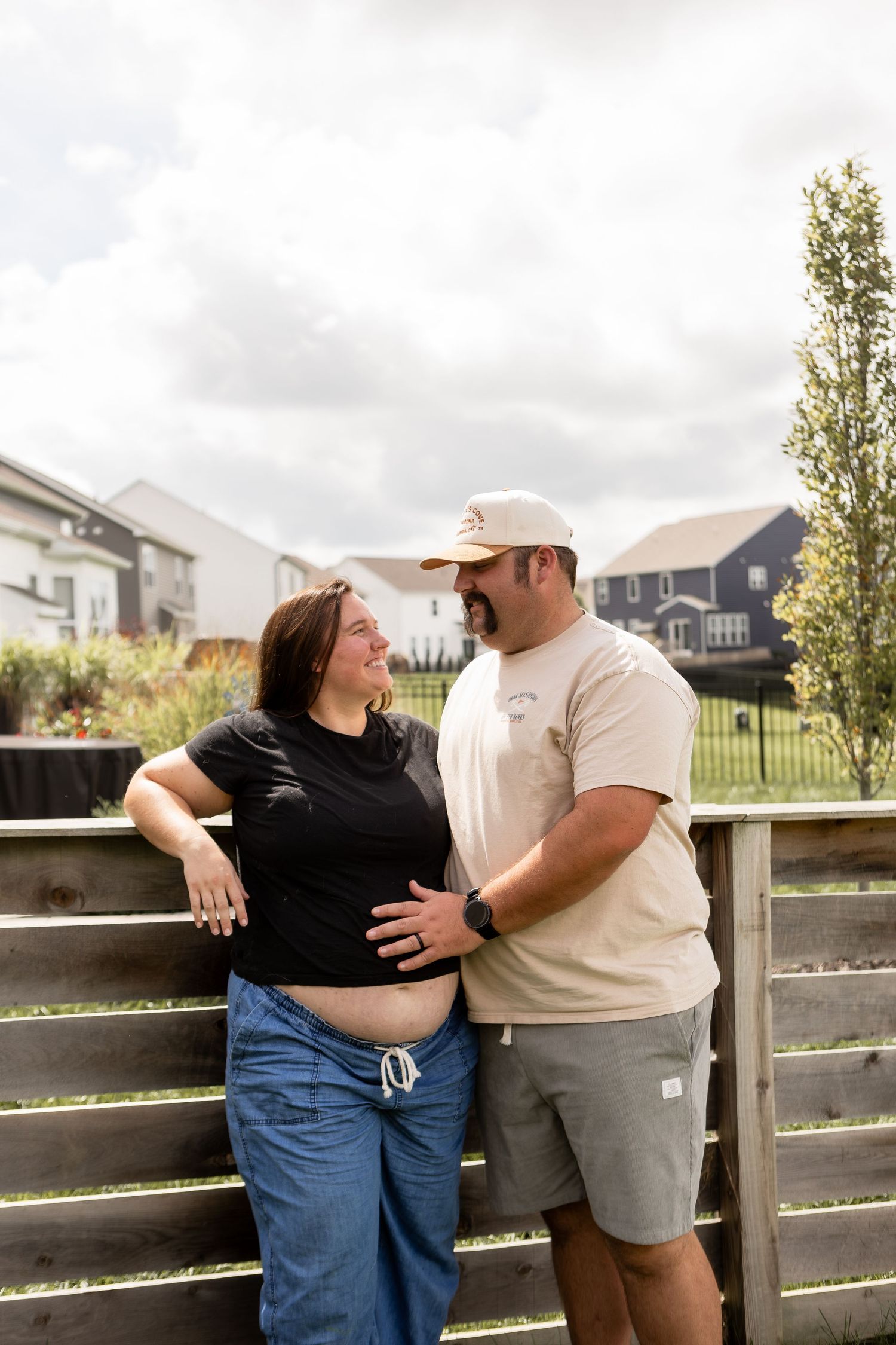 Pregnant woman leaning on fence with her husband embracing her belly at home for maternity session in Commercial Point Ohio