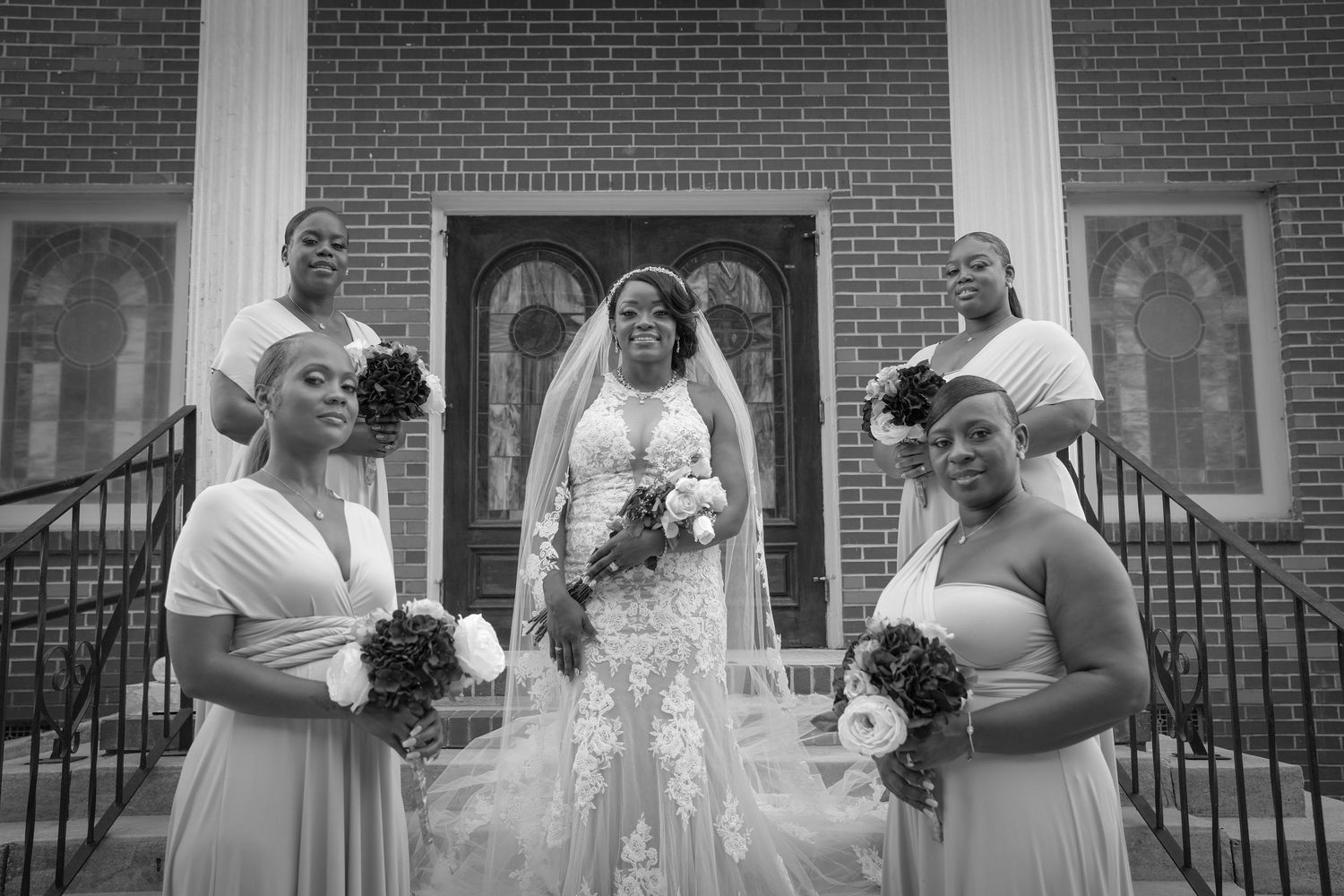 Black and white photo of a bride in lace gown with four bridesmaids holding bouquets on church steps.