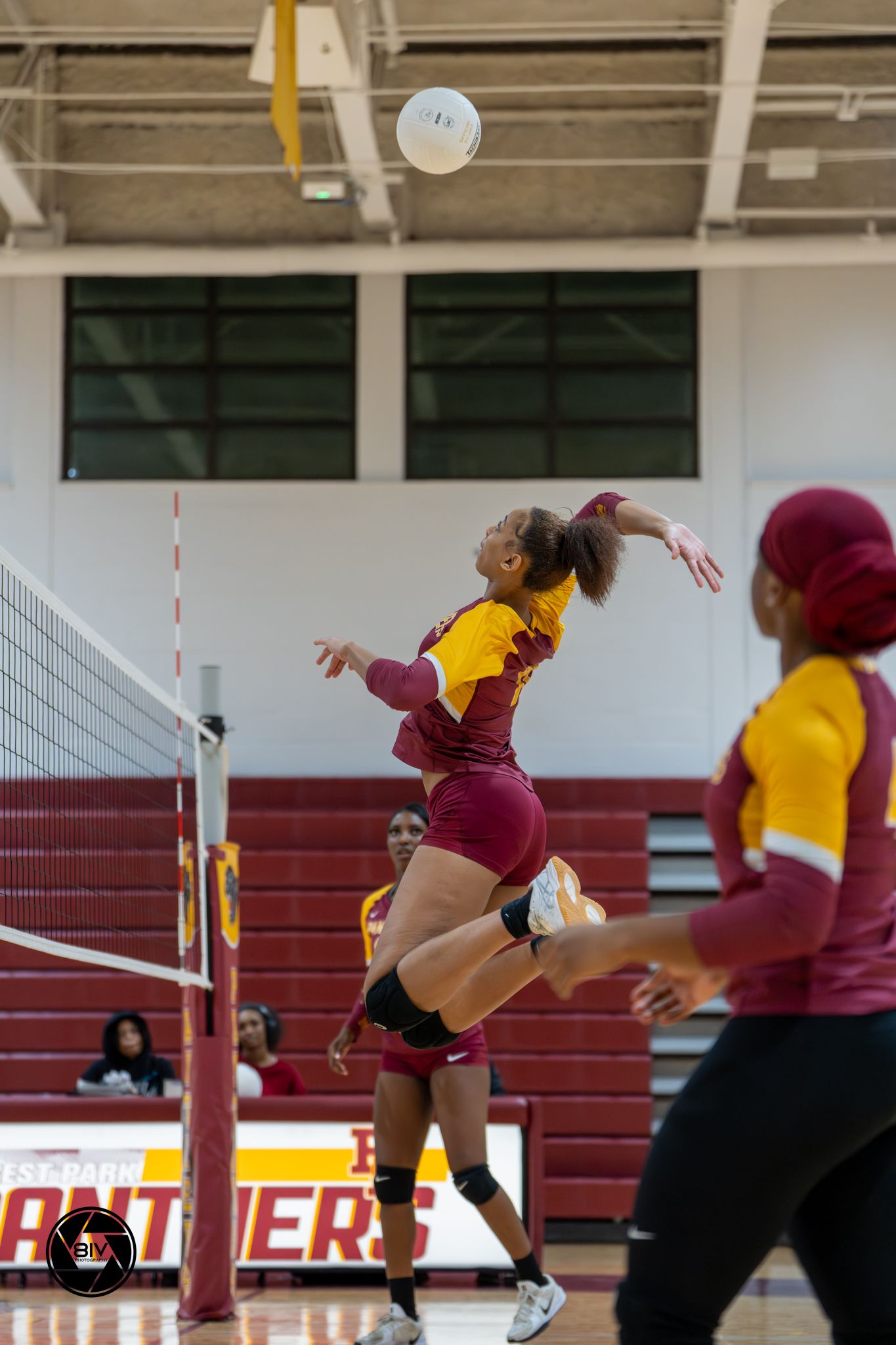 A female volleyball player in maroon and gold uniform jumps to spike the ball over the net during an indoor match.