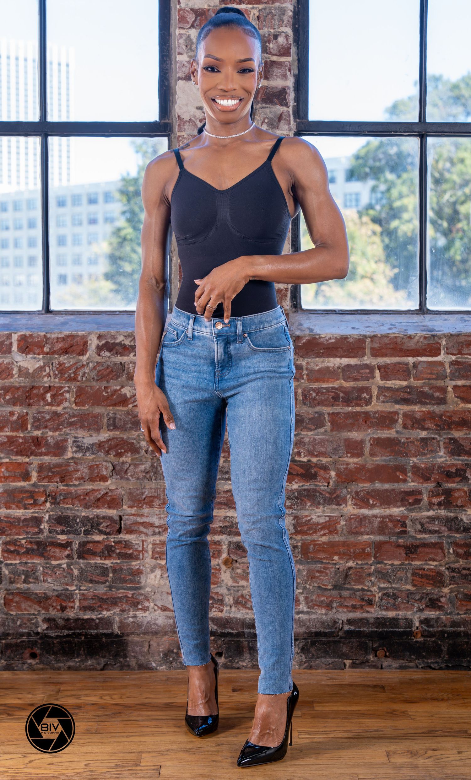 A tall woman in blue jeans and black bodysuit poses confidently in front of a brick wall near a window.