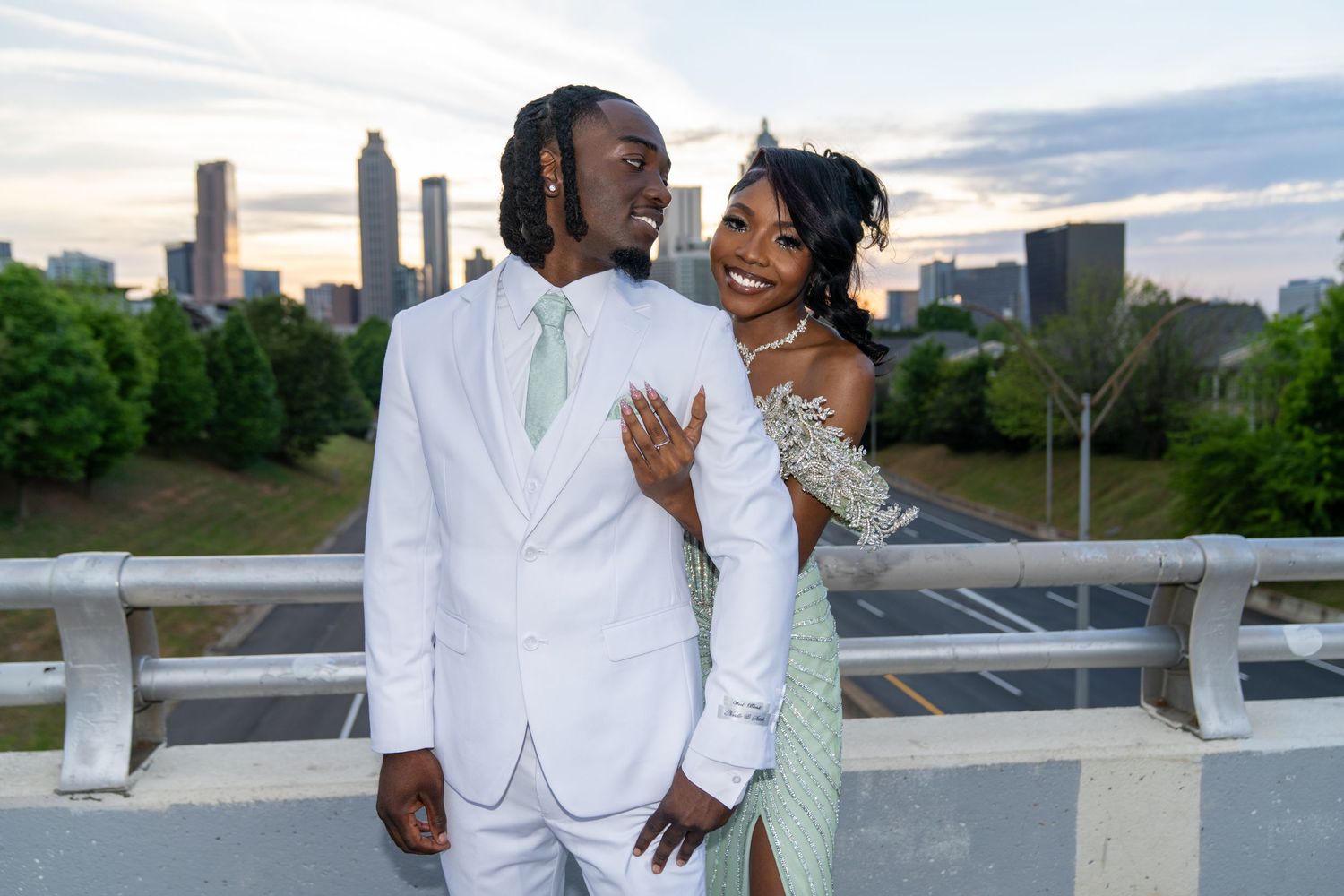 Couple in elegant prom attire posing on a bridge with Atlanta city skyline at sunset backdrop.