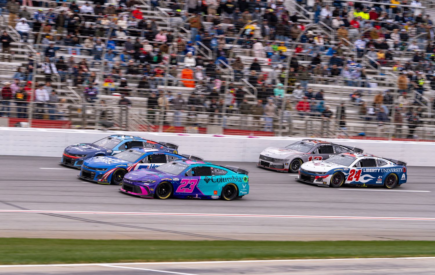 NASCAR race cars compete on a speedway track, featuring vibrant blue and pink liveries during an exciting multi-car race.