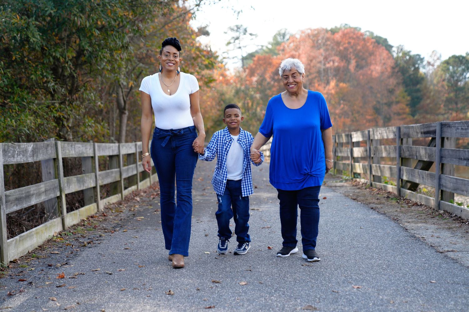 Three generations of women and child walking hand-in-hand on a scenic bridge path surrounded by autumn foliage.